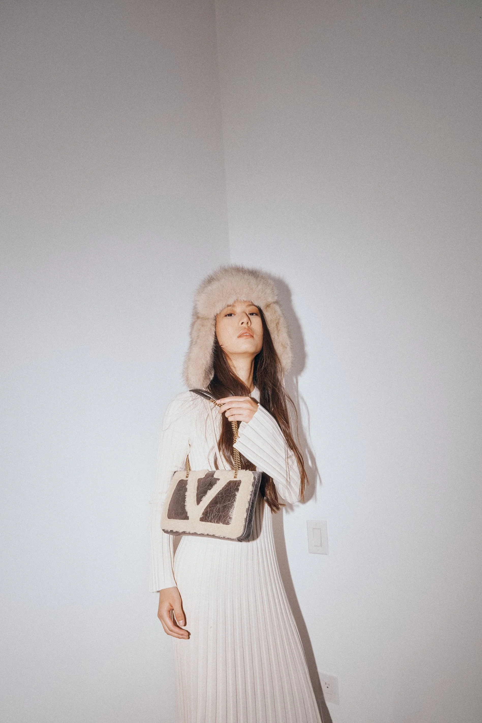 Woman in a cream pleated dress and pale furry hat holding a patterned handbag.
