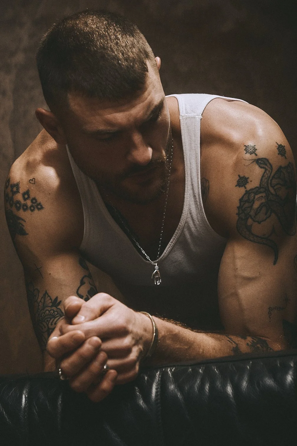 Close portrait of a tattooed man in a white tank top leaning forward with his hands clasped.