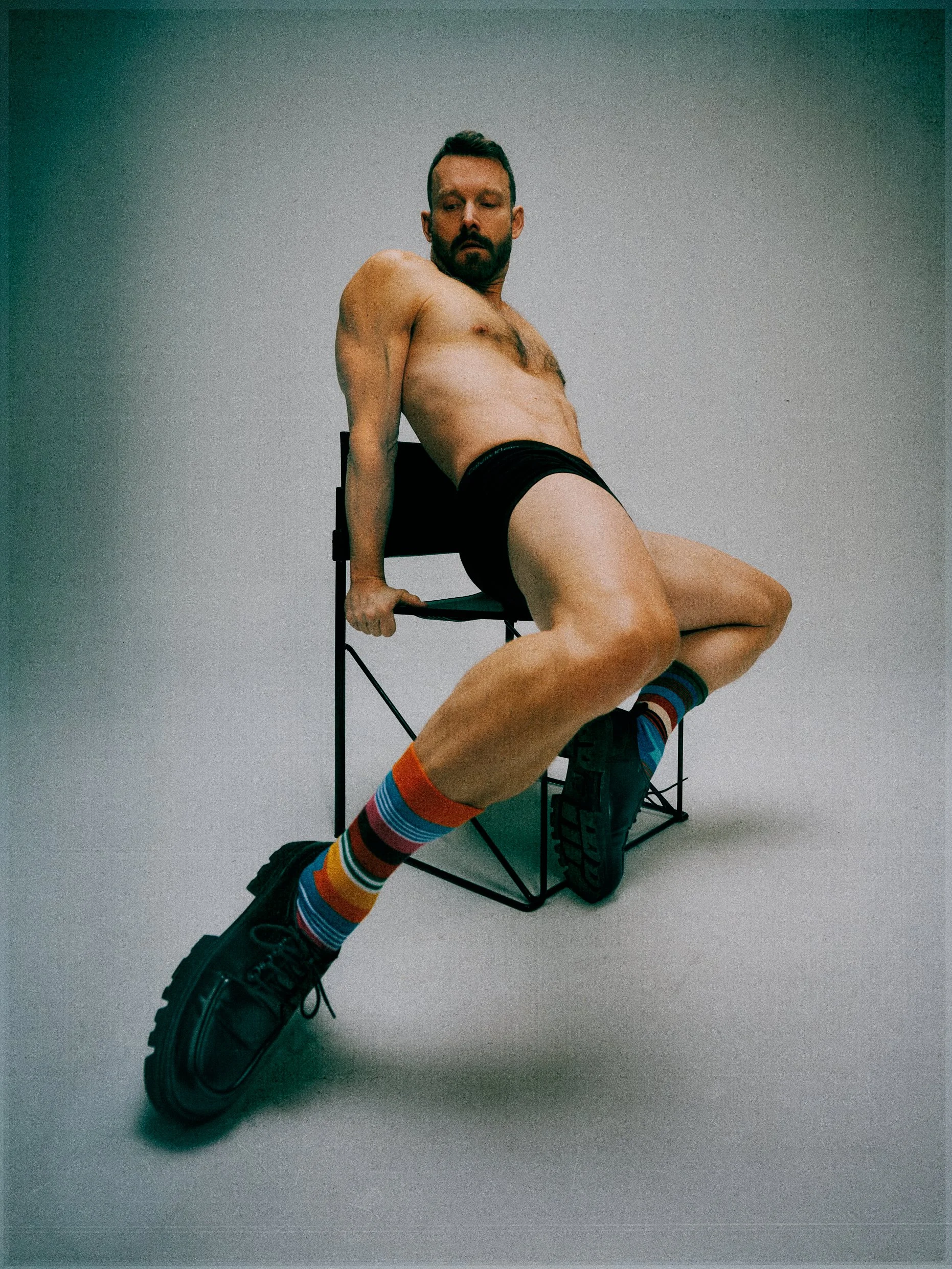 Shirtless man leaning back on a metal stool with crossed legs, striped socks and chunky shoes.