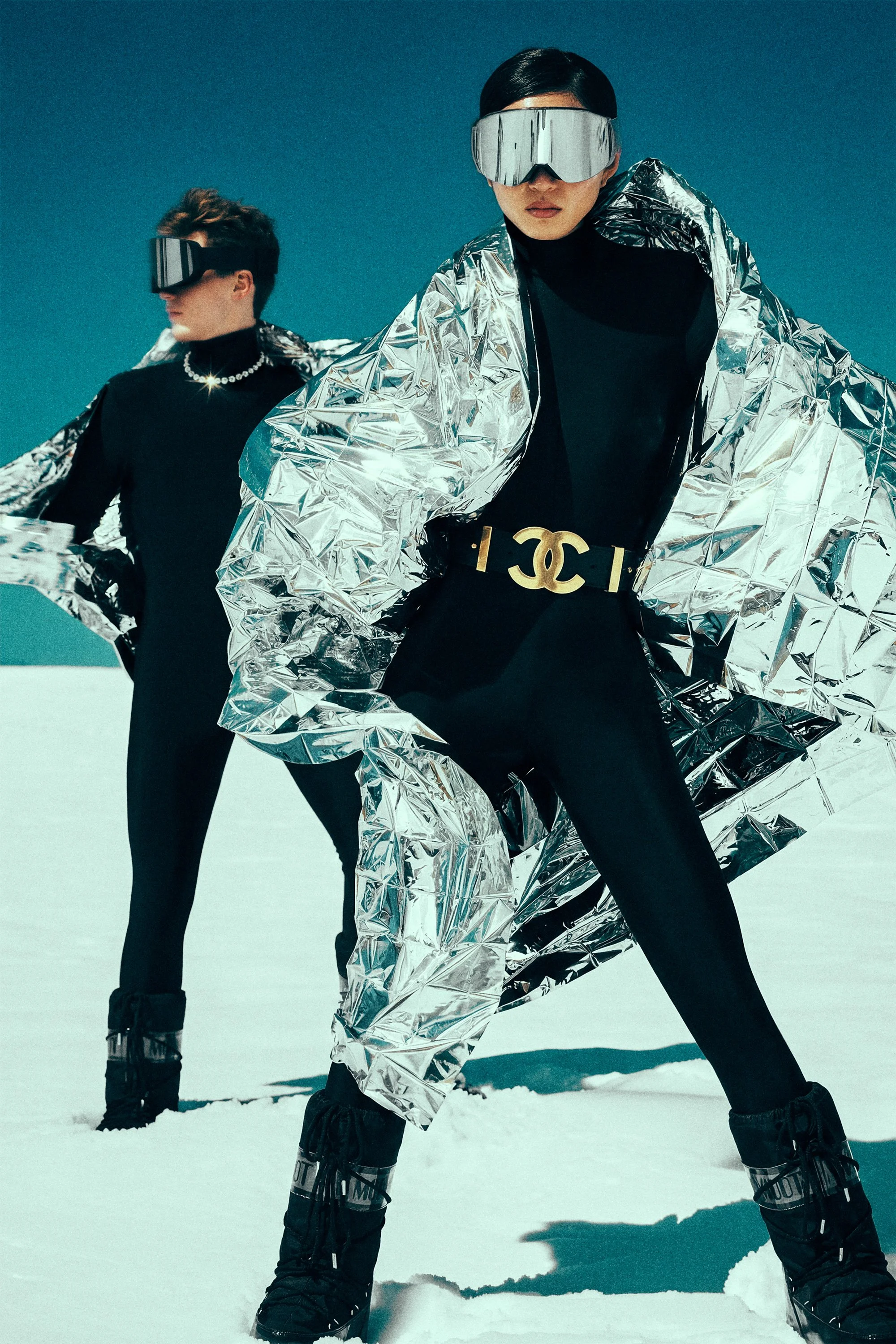 Two models in black outfits and goggles posing in snow, with silver outerwear in the foreground.