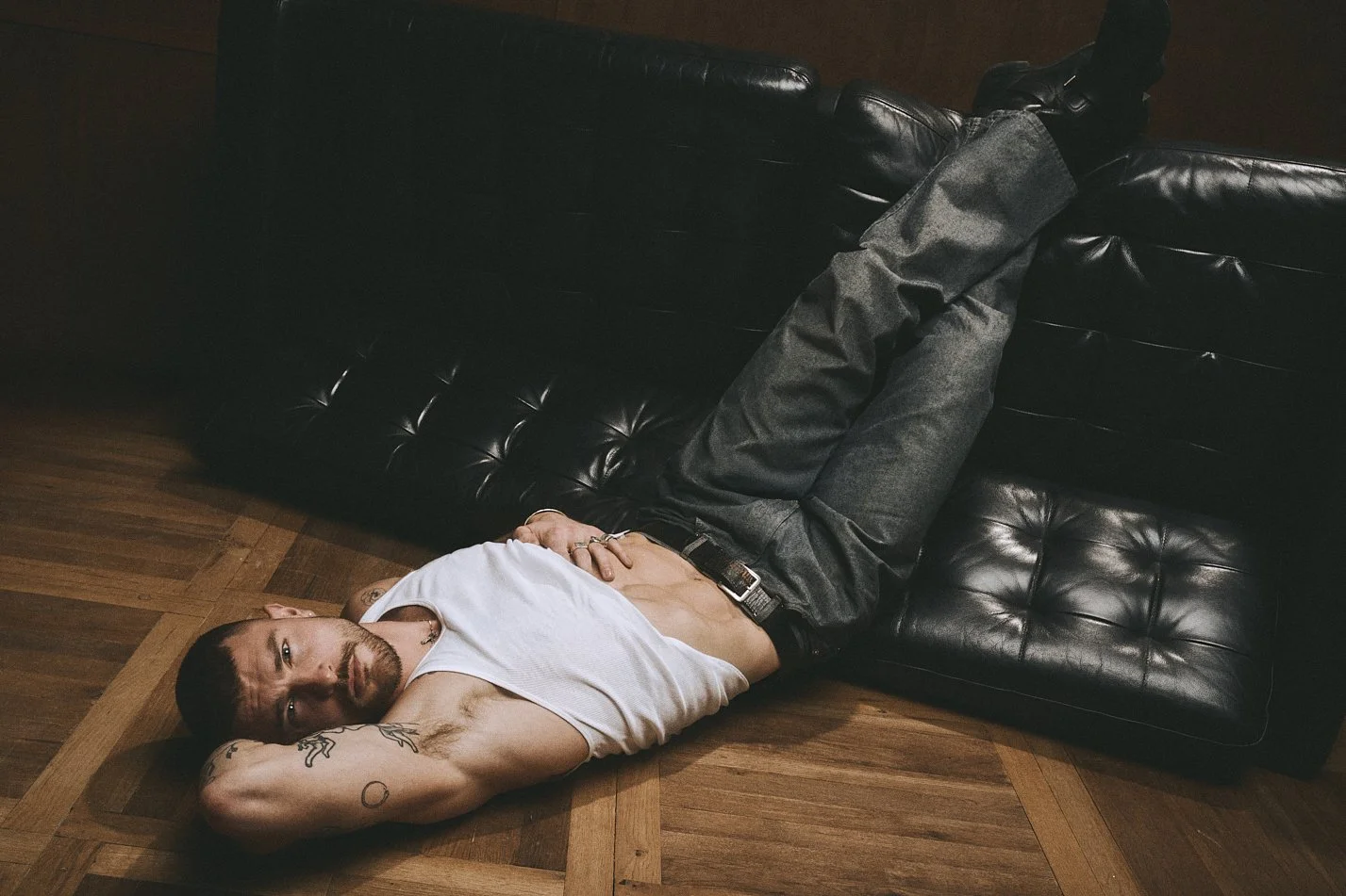 Tattooed man in a white tank top lying on a wooden floor with his legs resting on a leather sofa.