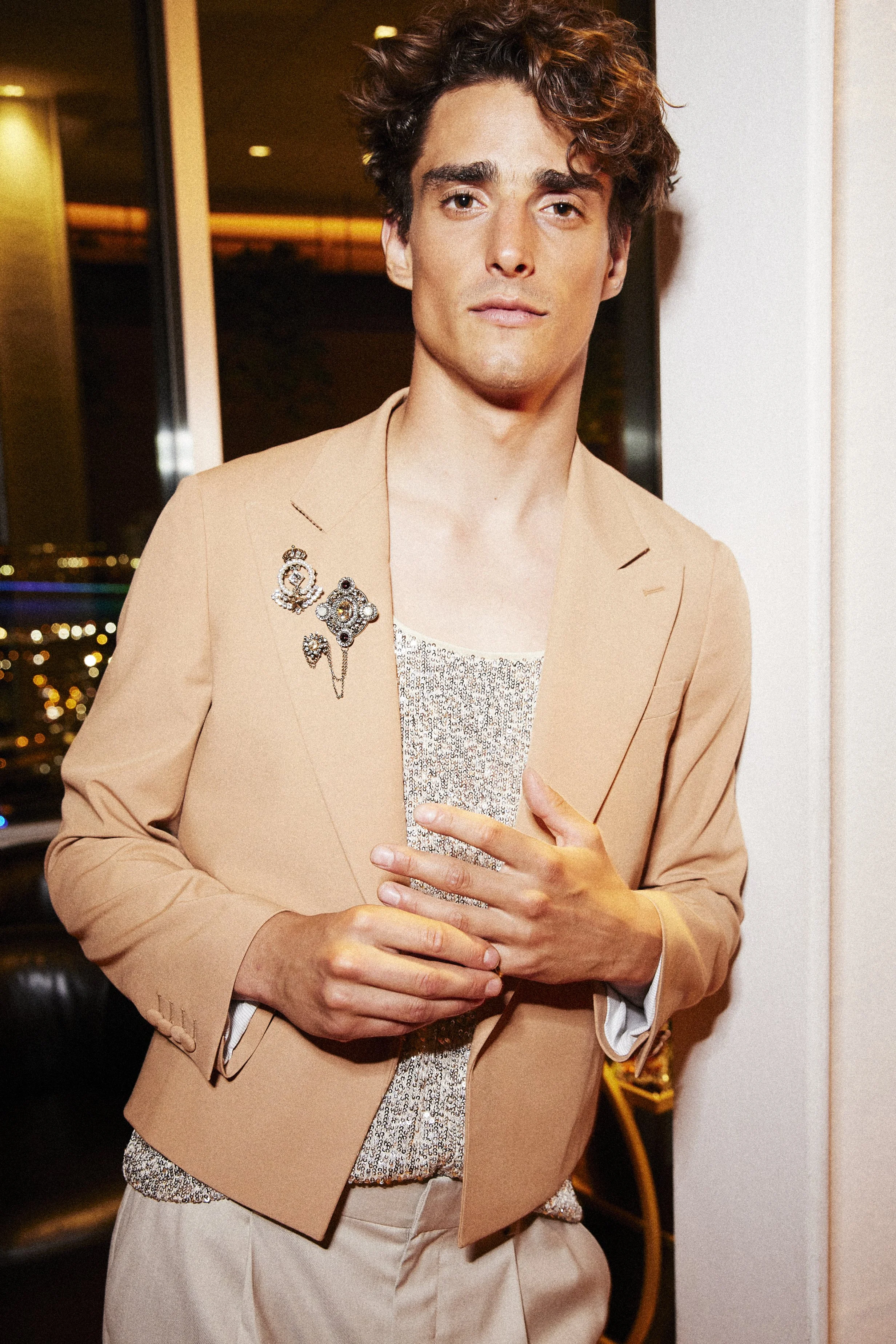 Man in a beige jacket over a sparkling top holding a drink.