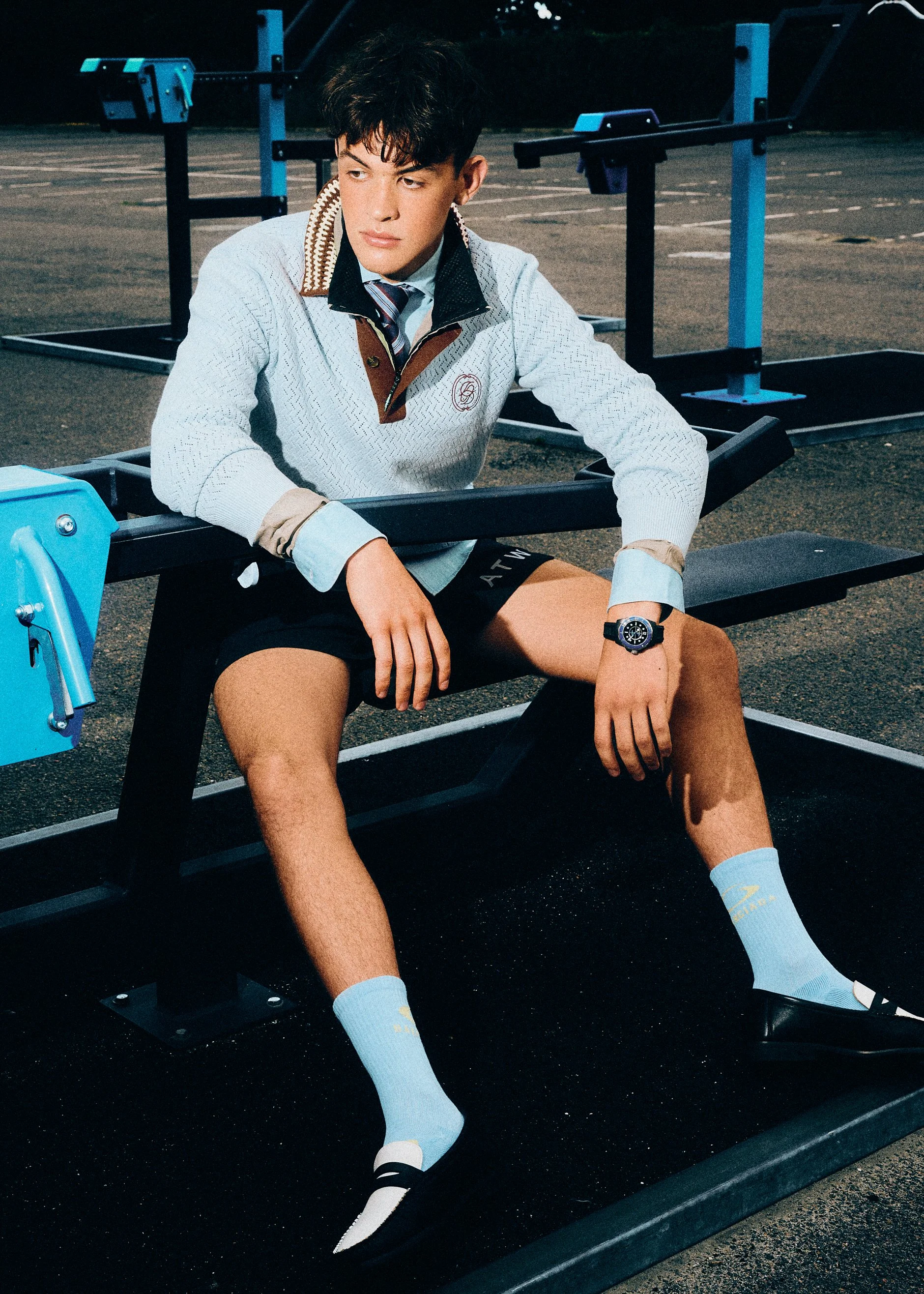 Man in a pale blue knit top, dark shorts and white socks seated on gym equipment.