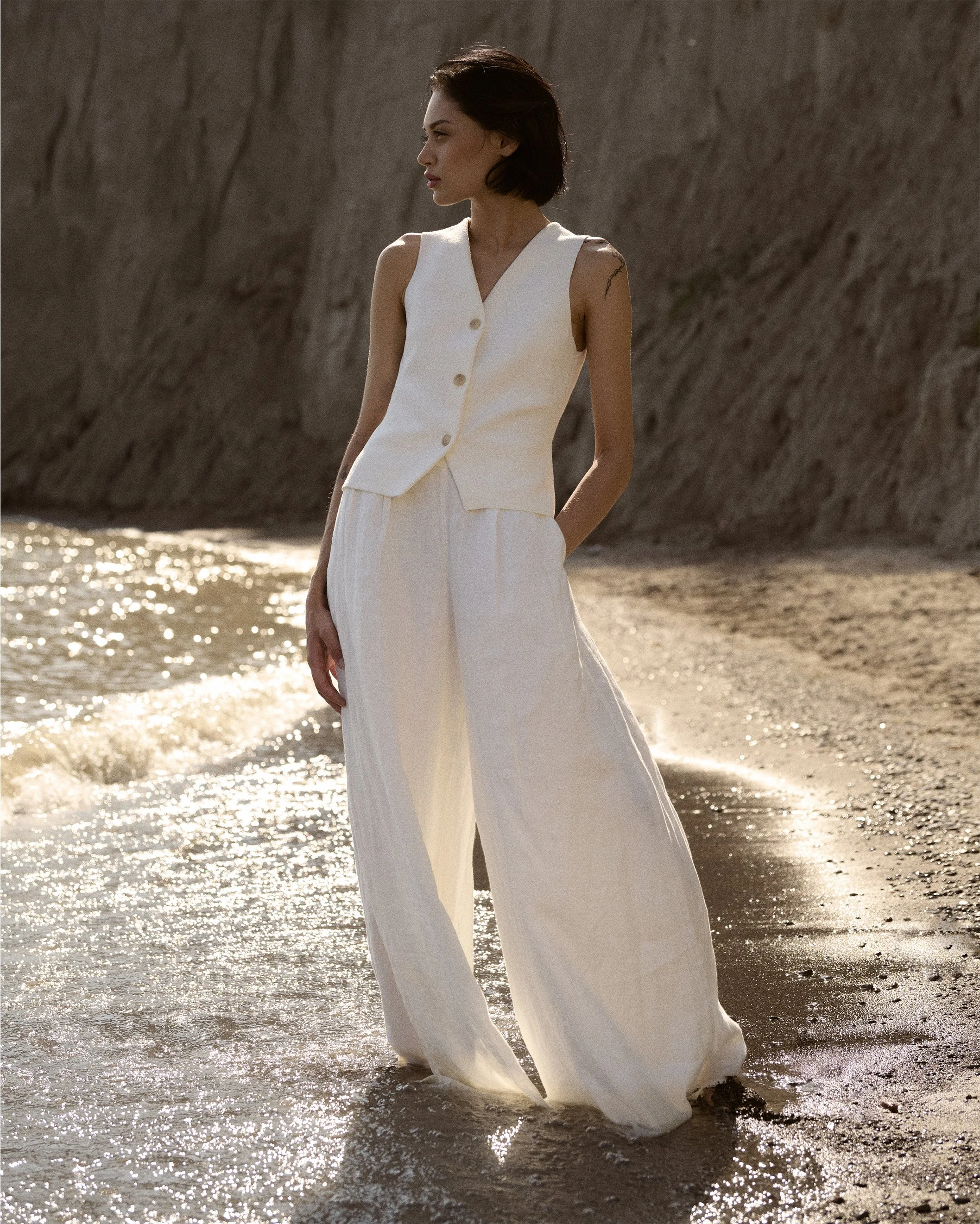 Woman in a white waistcoat and wide trousers standing beside sunlit water and a rocky cliff.