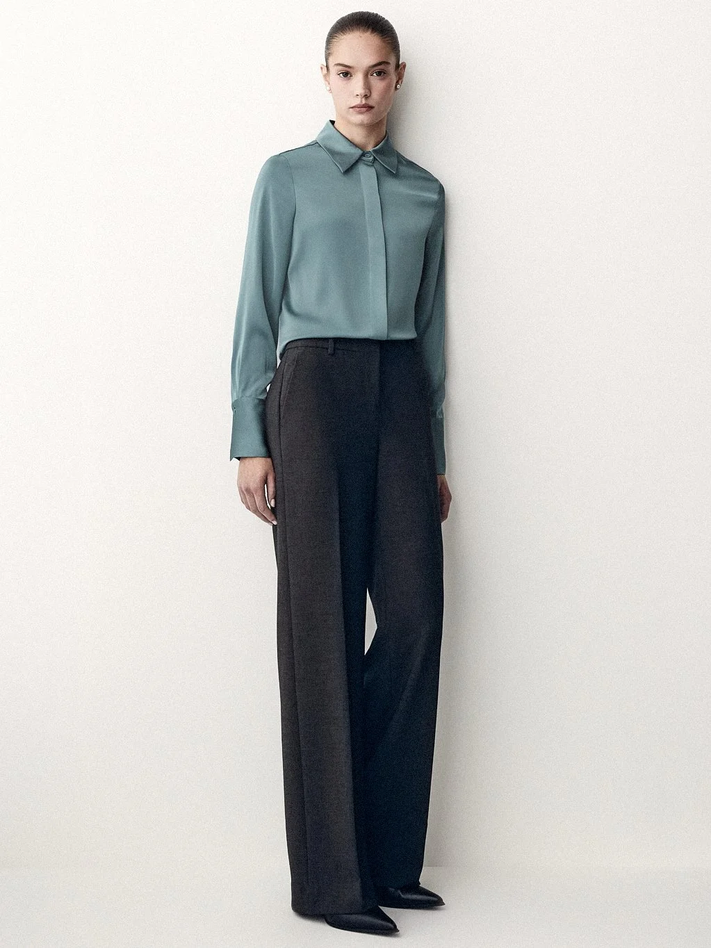 Woman in a muted teal blouse with a neck tie and wide dark trousers.