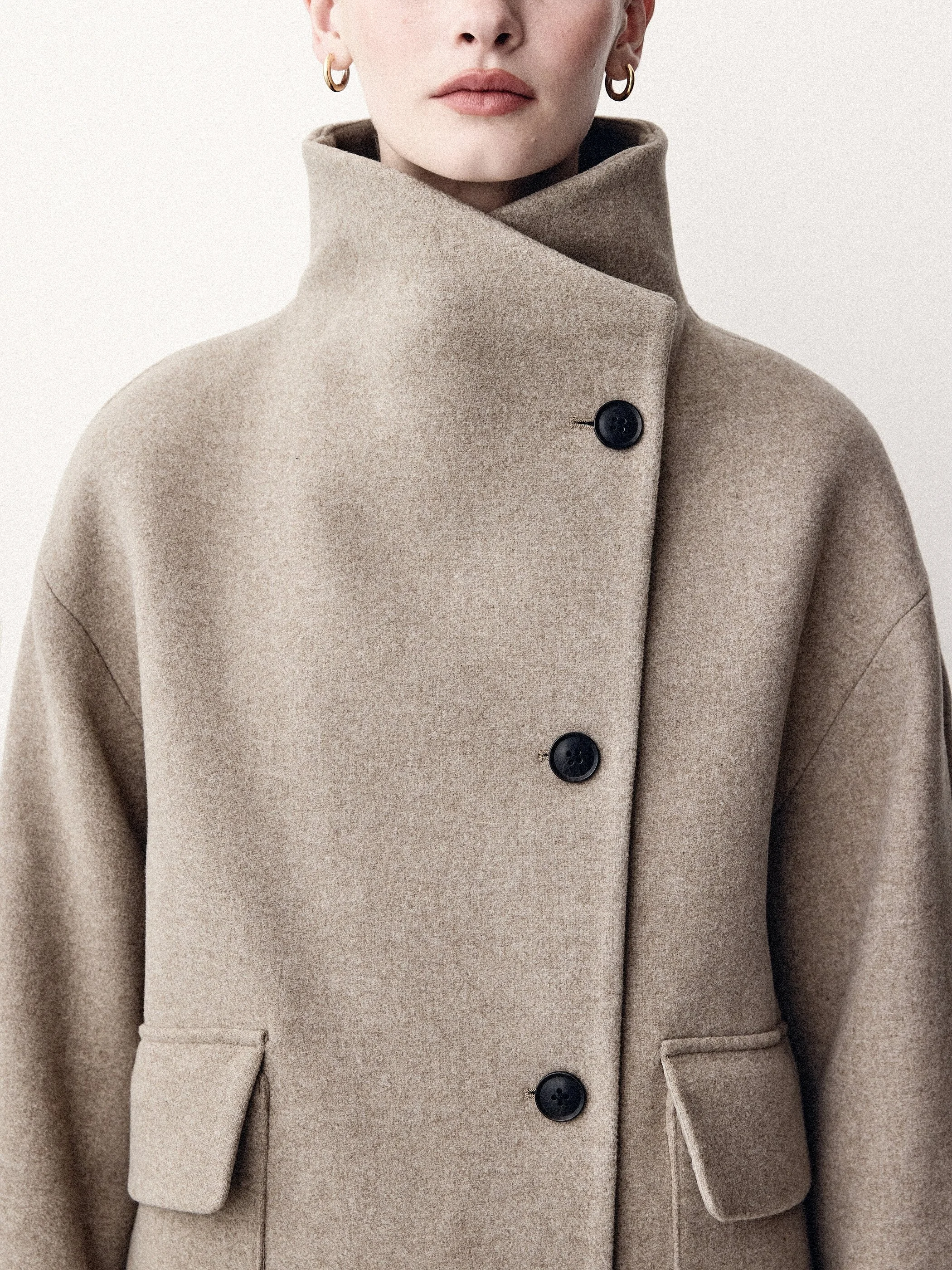 Detail of a taupe coat with an asymmetric high collar and large dark buttons.