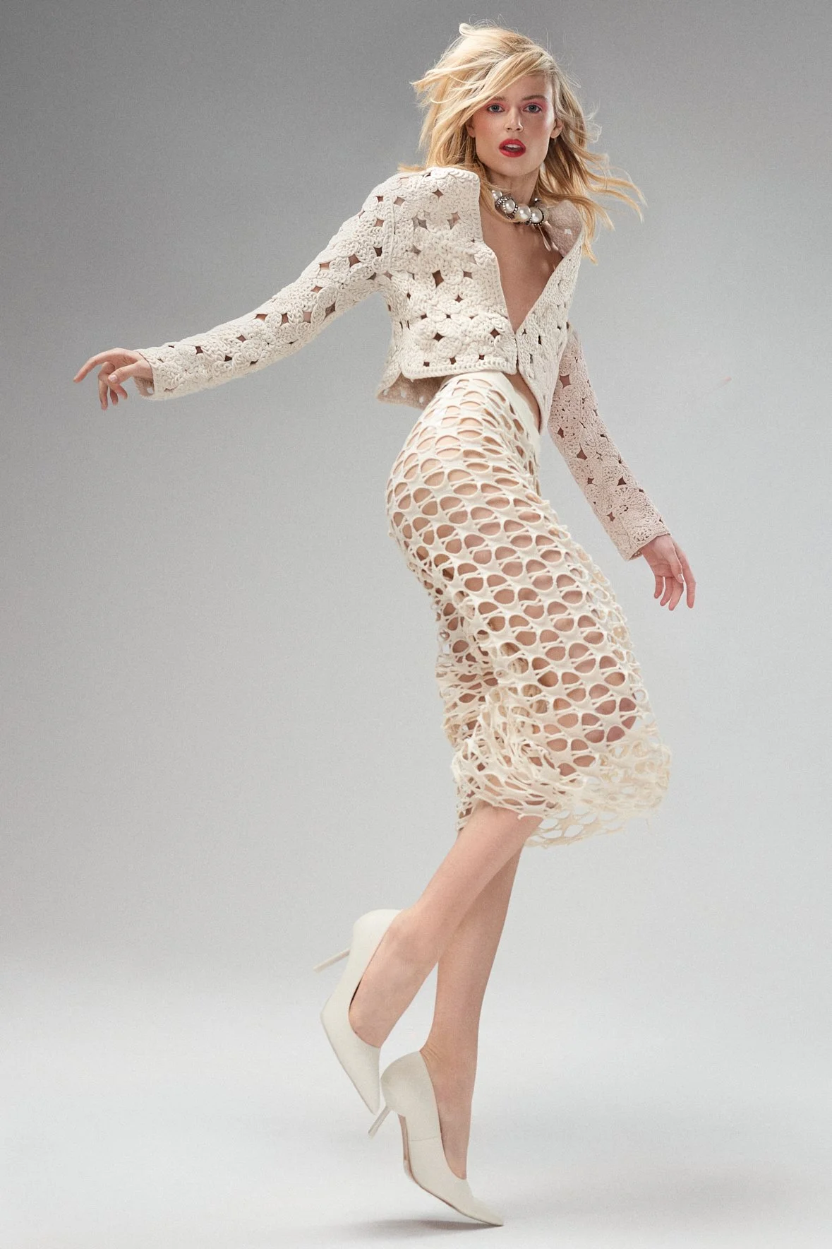 Blonde woman moving in a cream crochet jacket and matching skirt against a grey studio background.