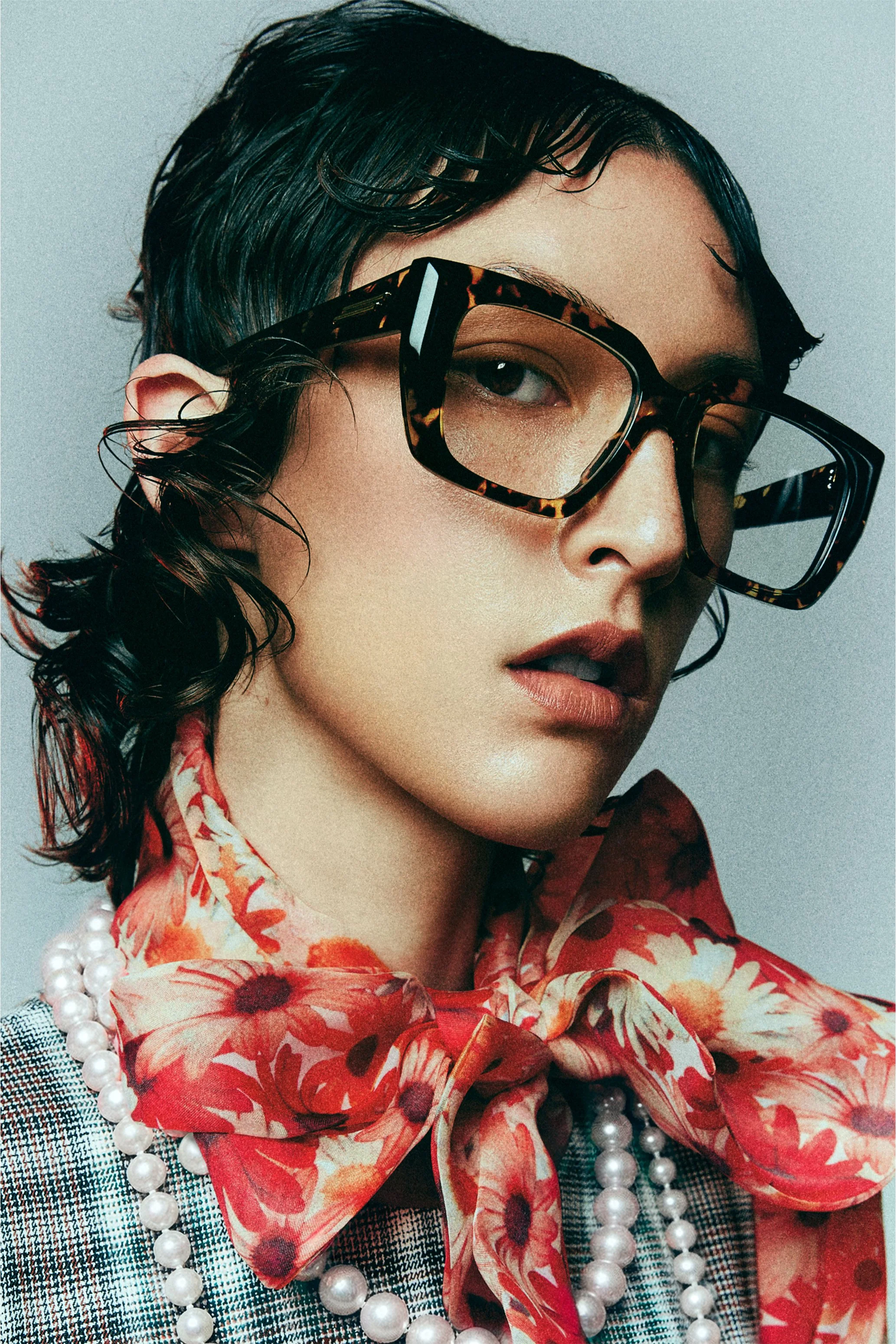Beauty close-up of a woman in oversized glasses, a floral neck scarf and layered pearl necklaces.