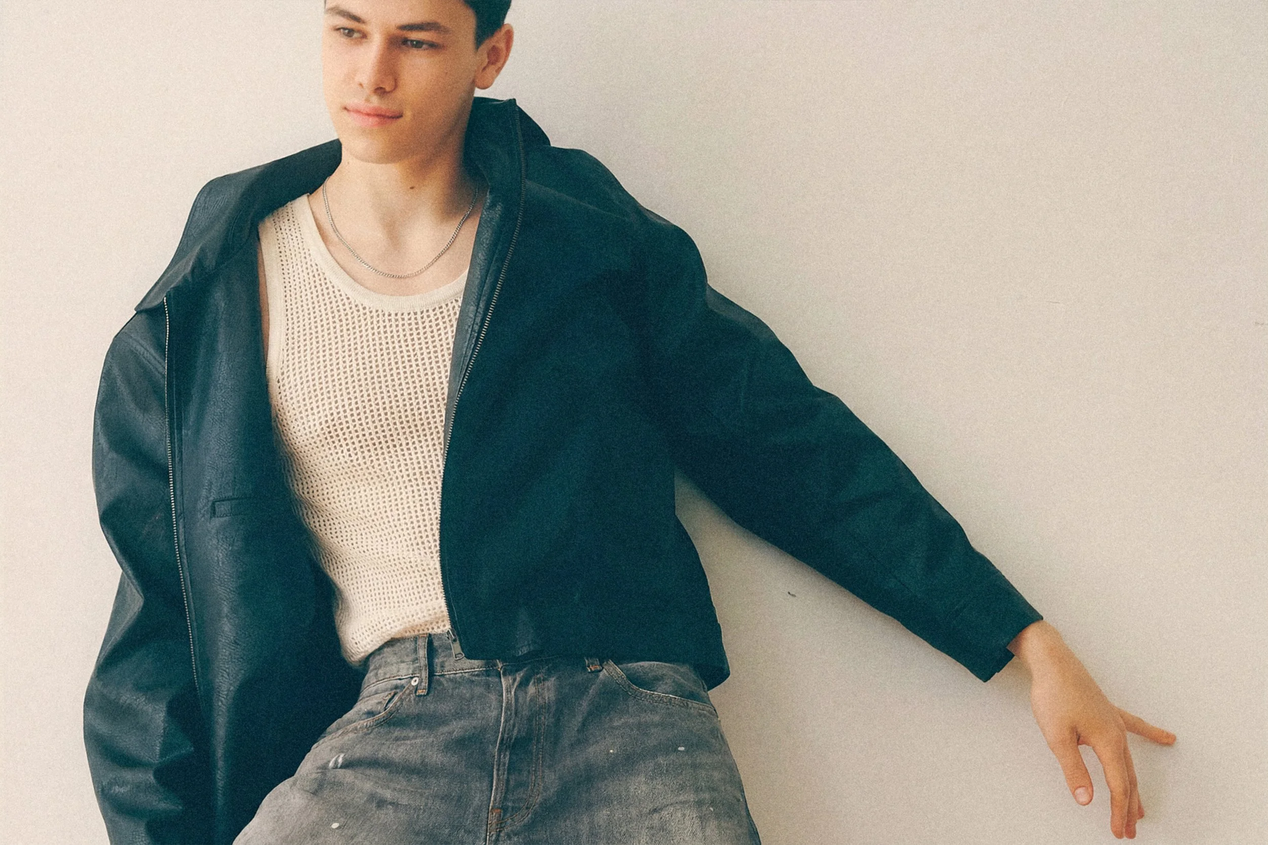 Closer view of a young man leaning against a wall in a black jacket over a white tank top.