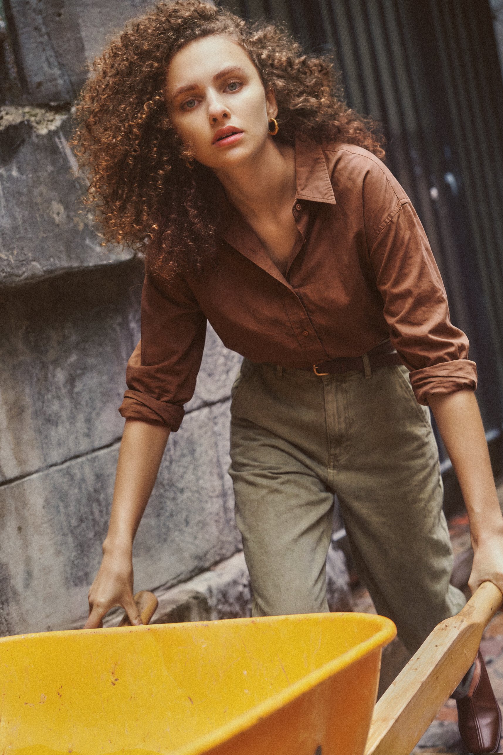 Fashion model in a brown shirt leaning forward over a yellow wheelbarrow, photographed in a gritty urban setting.