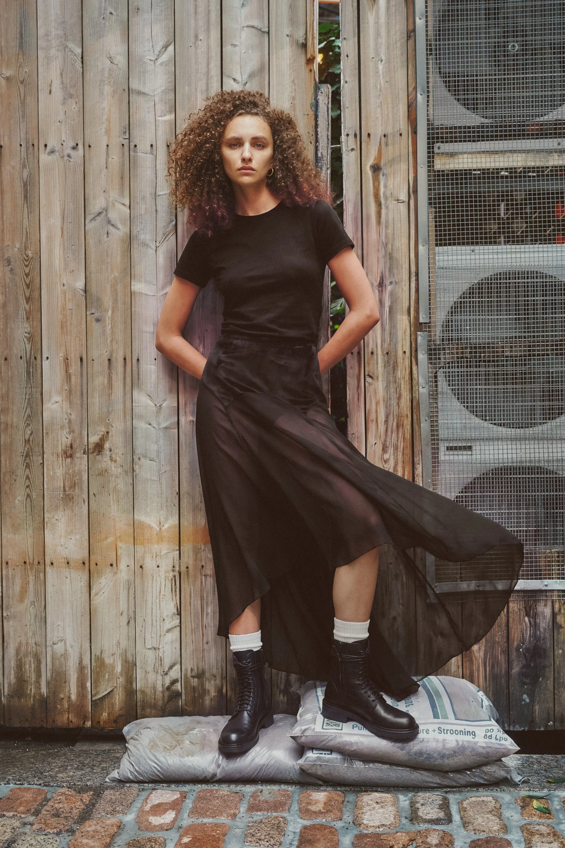 Curly-haired model wearing a black T-shirt, flowing sheer skirt and combat boots against a wooden wall in an urban courtyard.