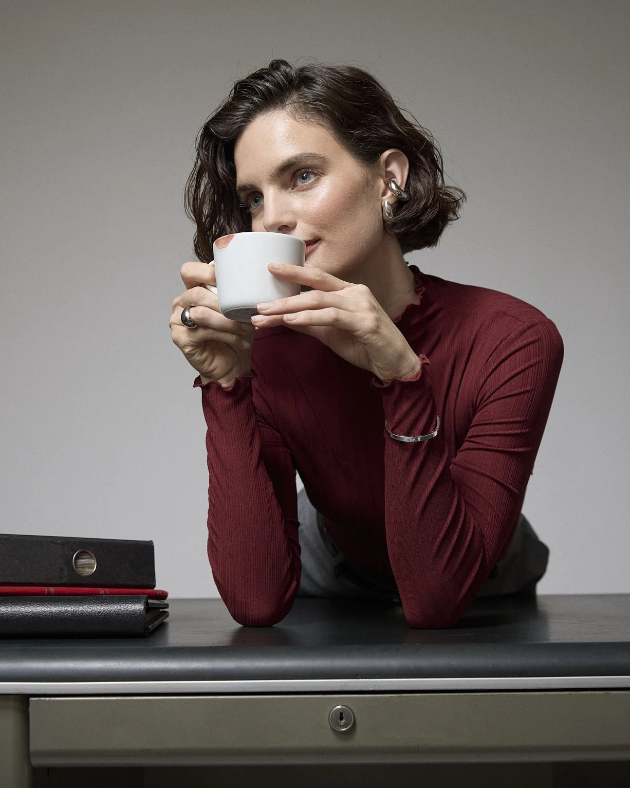 Woman in a burgundy blouse holding a white cup while leaning on an office desk.