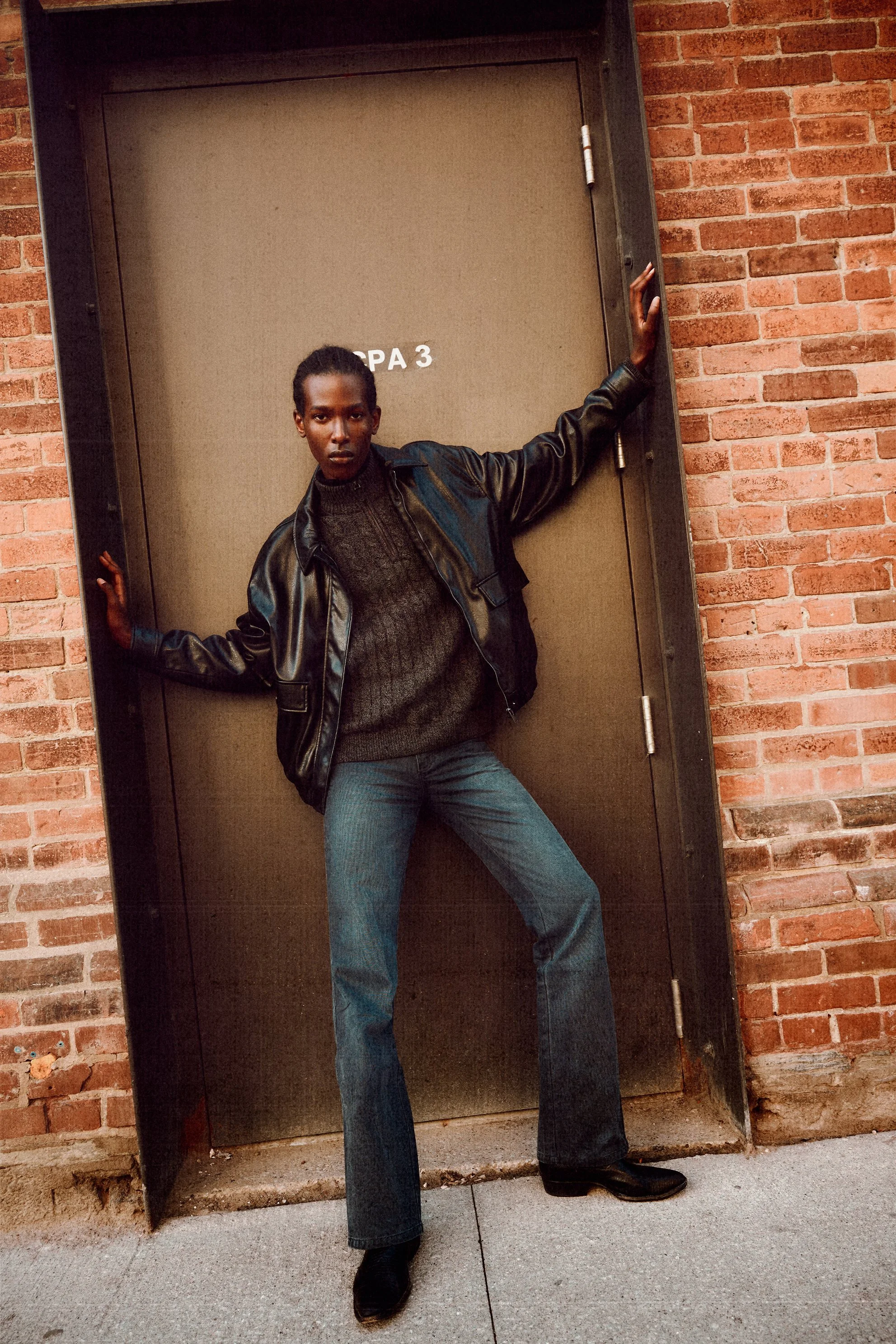 Man in a leather jacket and jeans standing with his arms spread across an industrial doorway.