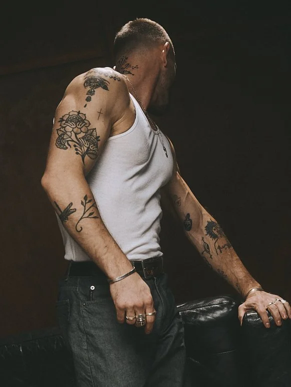 Tattooed man in a white tank top and dark trousers turning away with one hand on a leather sofa.