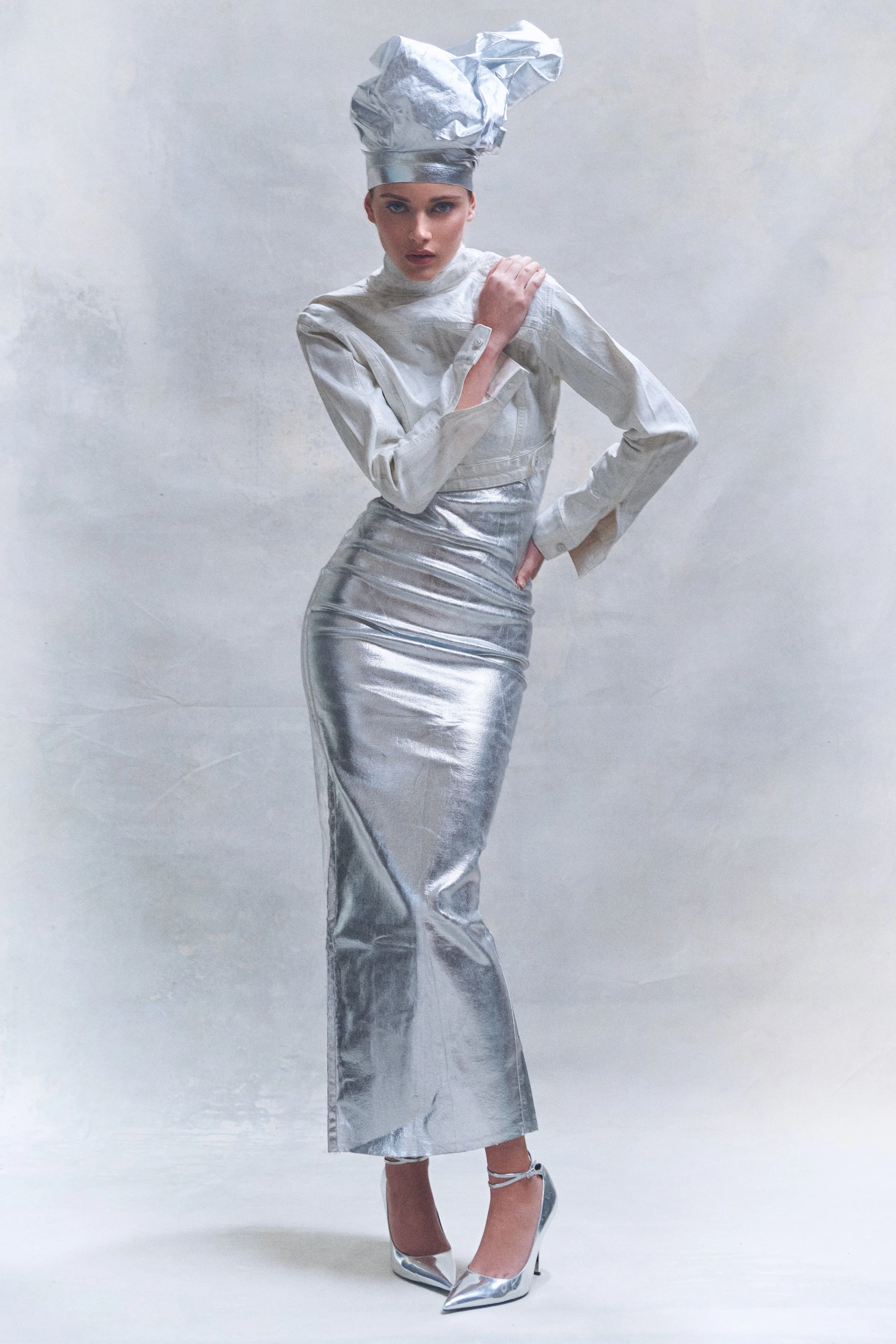 Woman in a sculptural silver dress and matching oversized headpiece, with one hand on her hip.