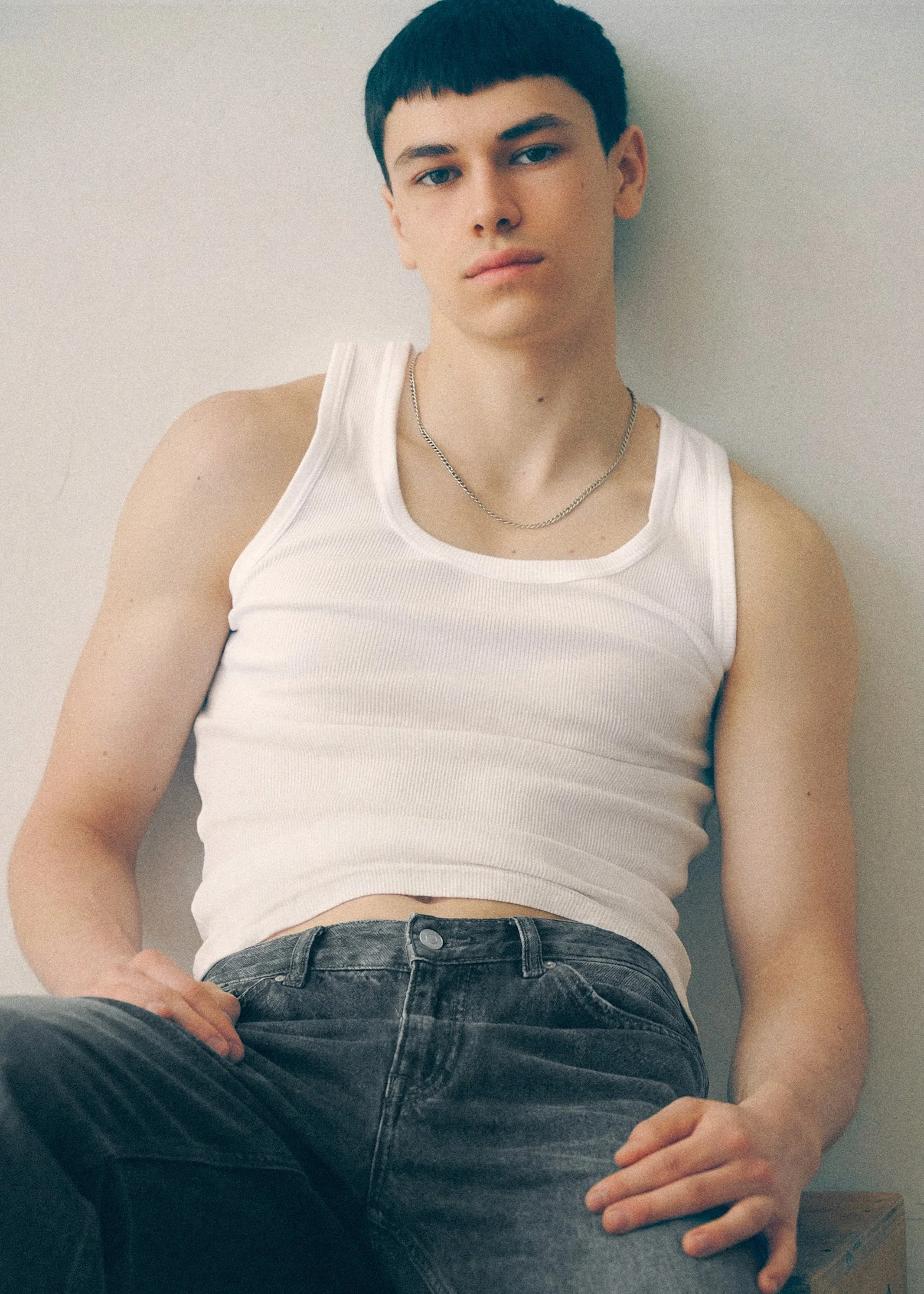 Seated portrait of a young man in a white tank top and jeans looking directly at the camera.