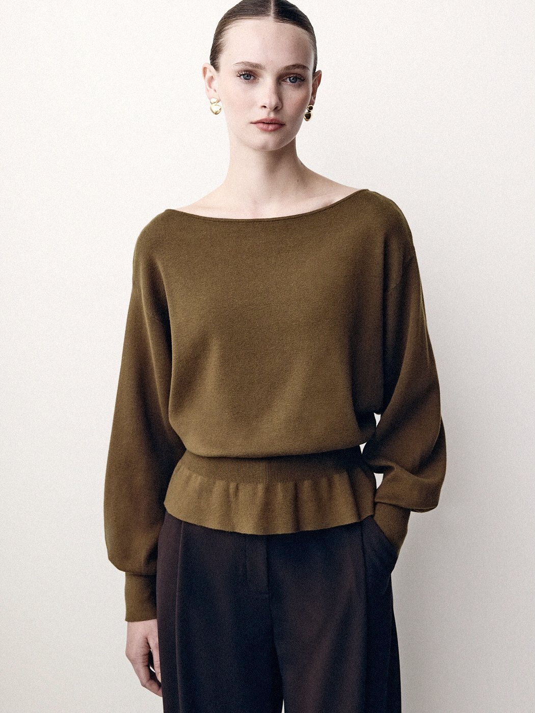 Woman wearing a brown belted sweater and dark trousers, hands in her pockets.