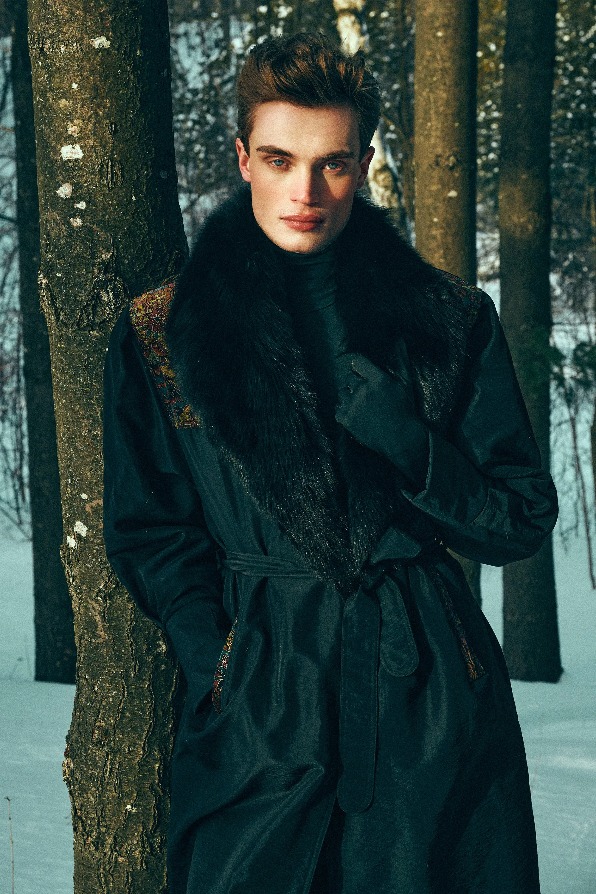 Man in a long black coat standing among trees in a snowy forest.