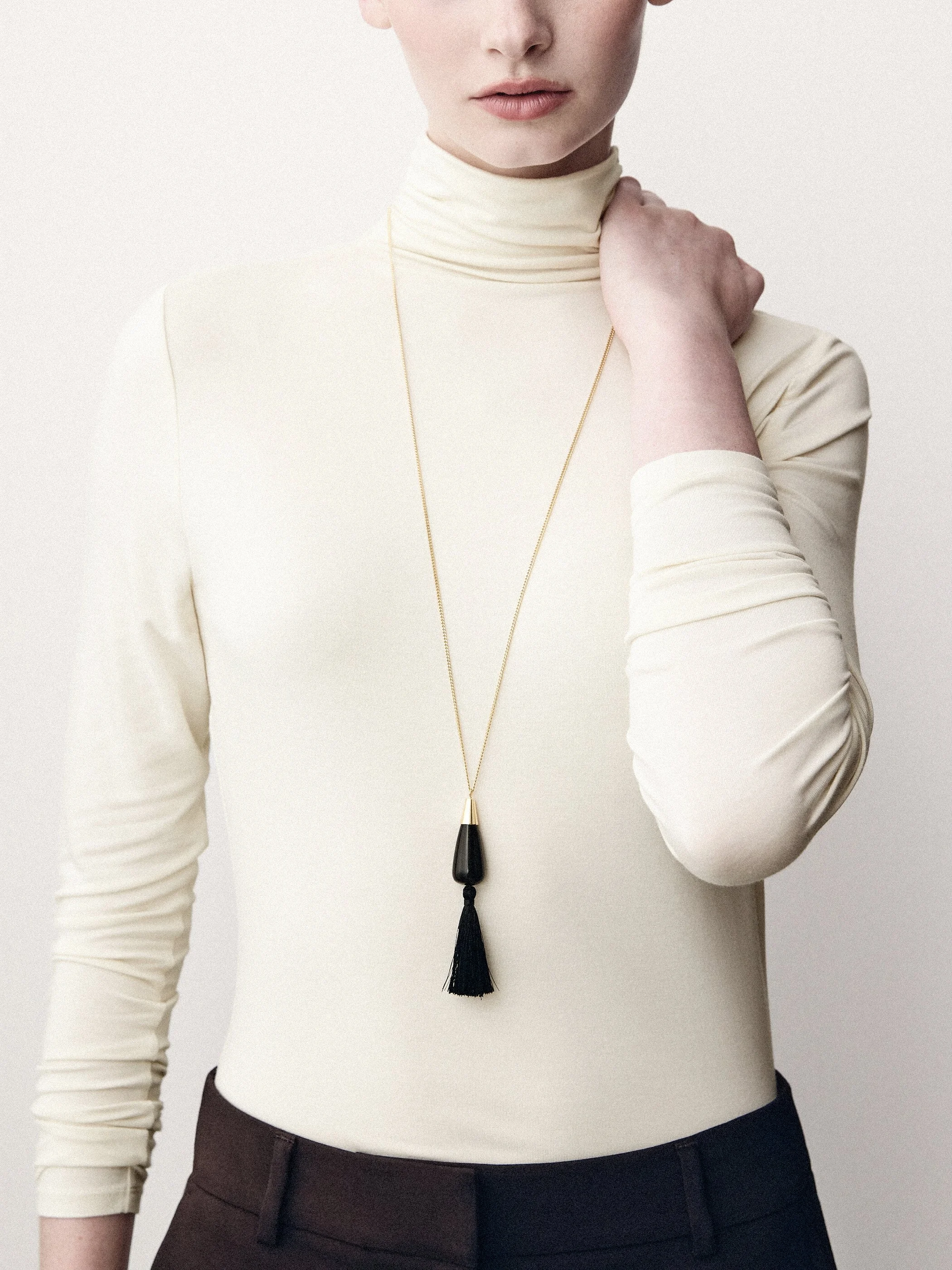 Detail of a cream turtleneck and long tassel necklace, with one hand raised to the collar.