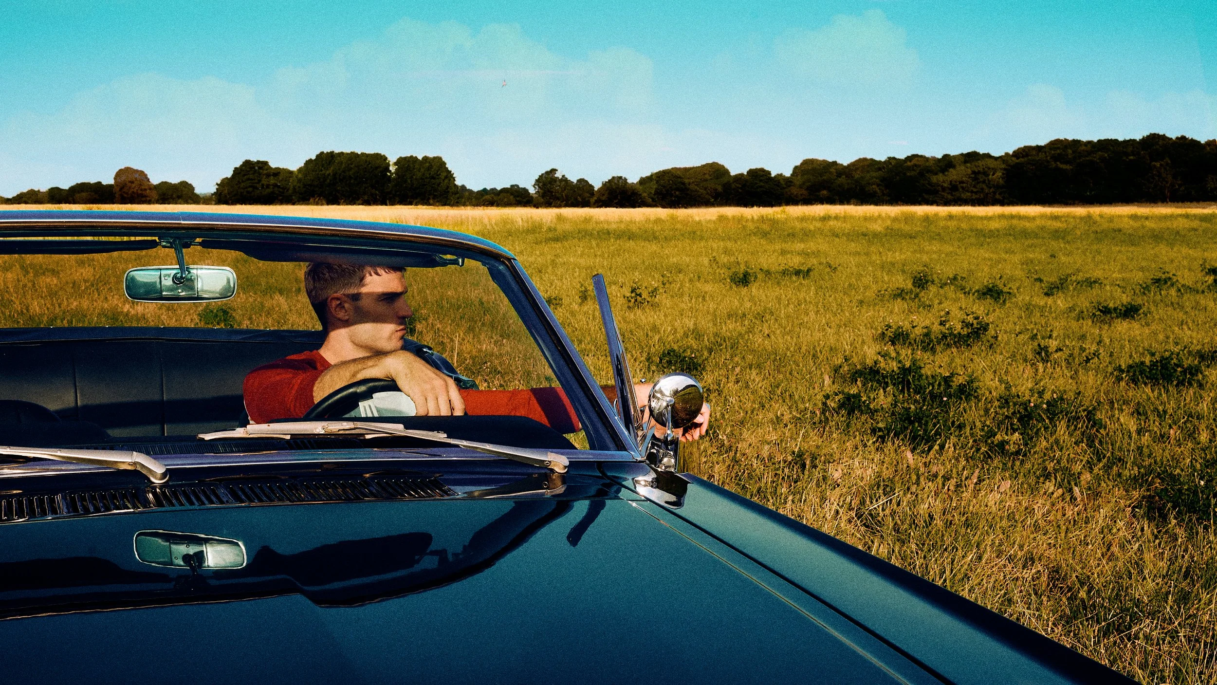 Man seated behind the windshield of a turquoise convertible in a sunlit field.