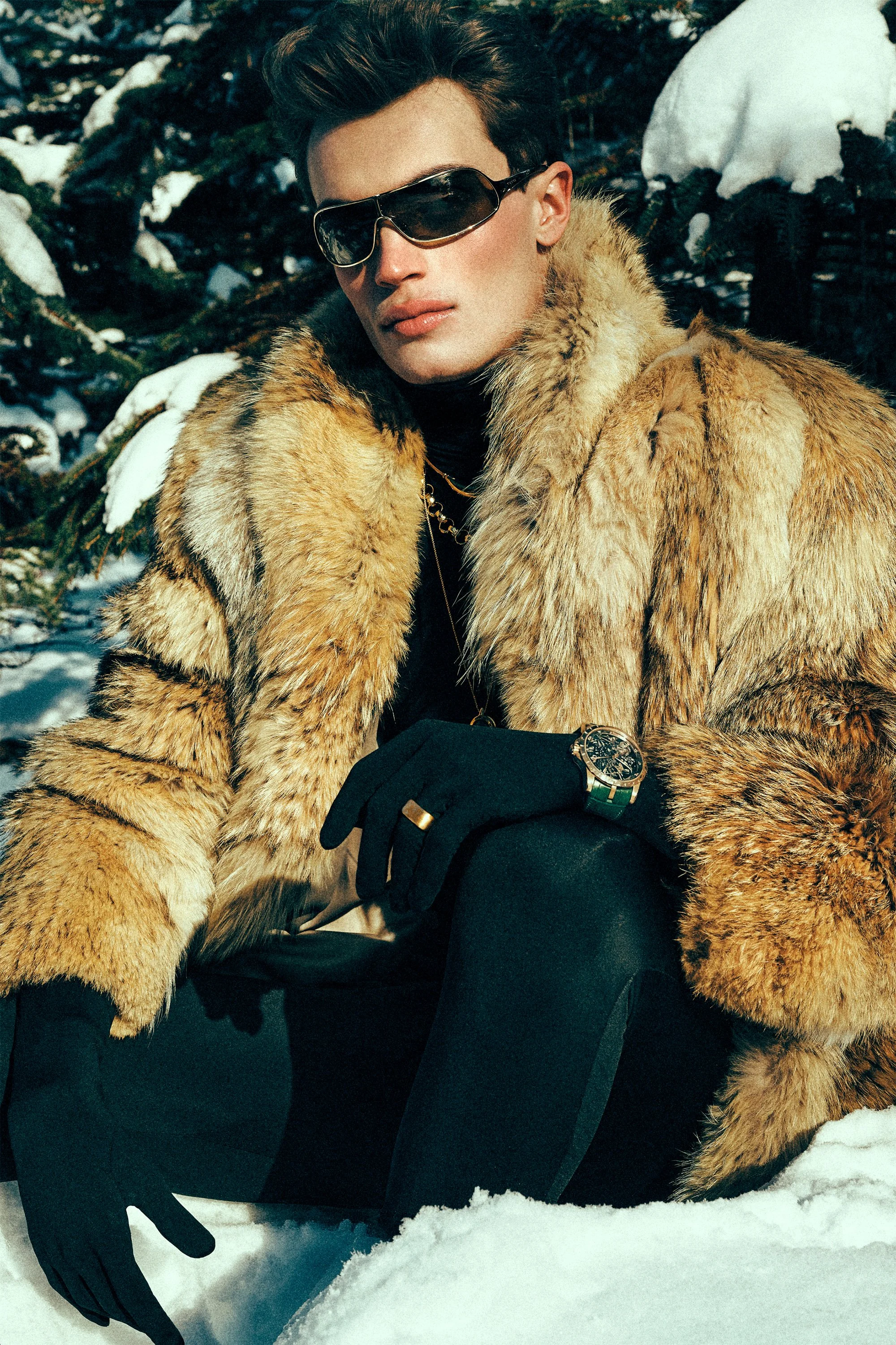 Man in sunglasses, black gloves and a brown fur-textured coat beside snowy branches.