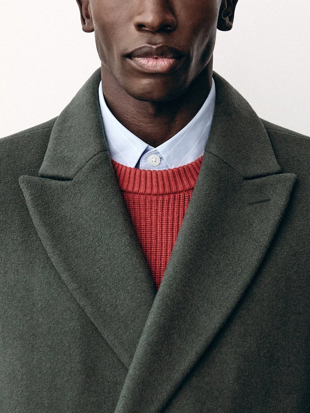 Close crop of an olive coat layered over a red sweater and blue collared shirt.