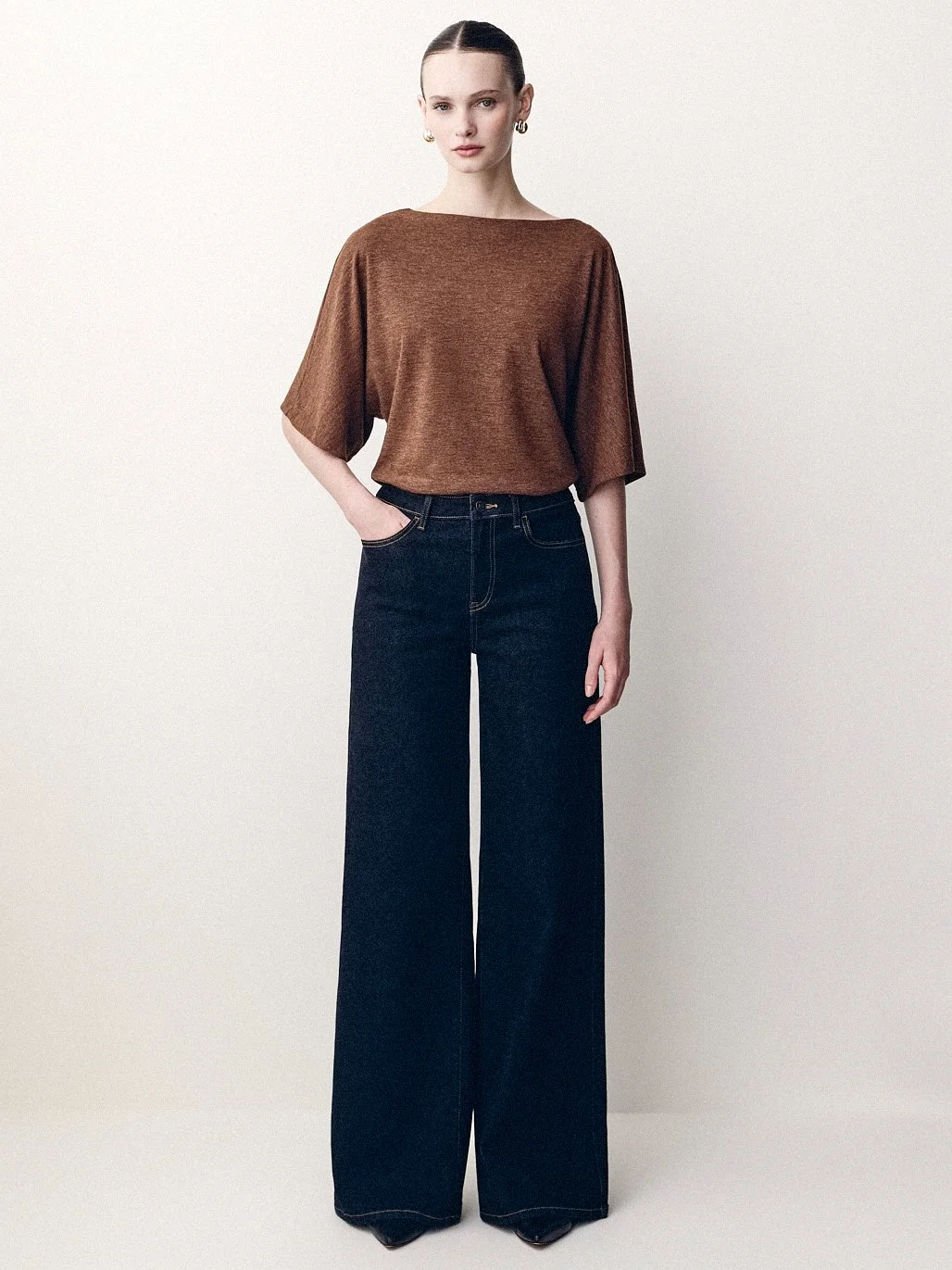 Woman in a loose brown knit top and wide dark jeans standing in the studio.
