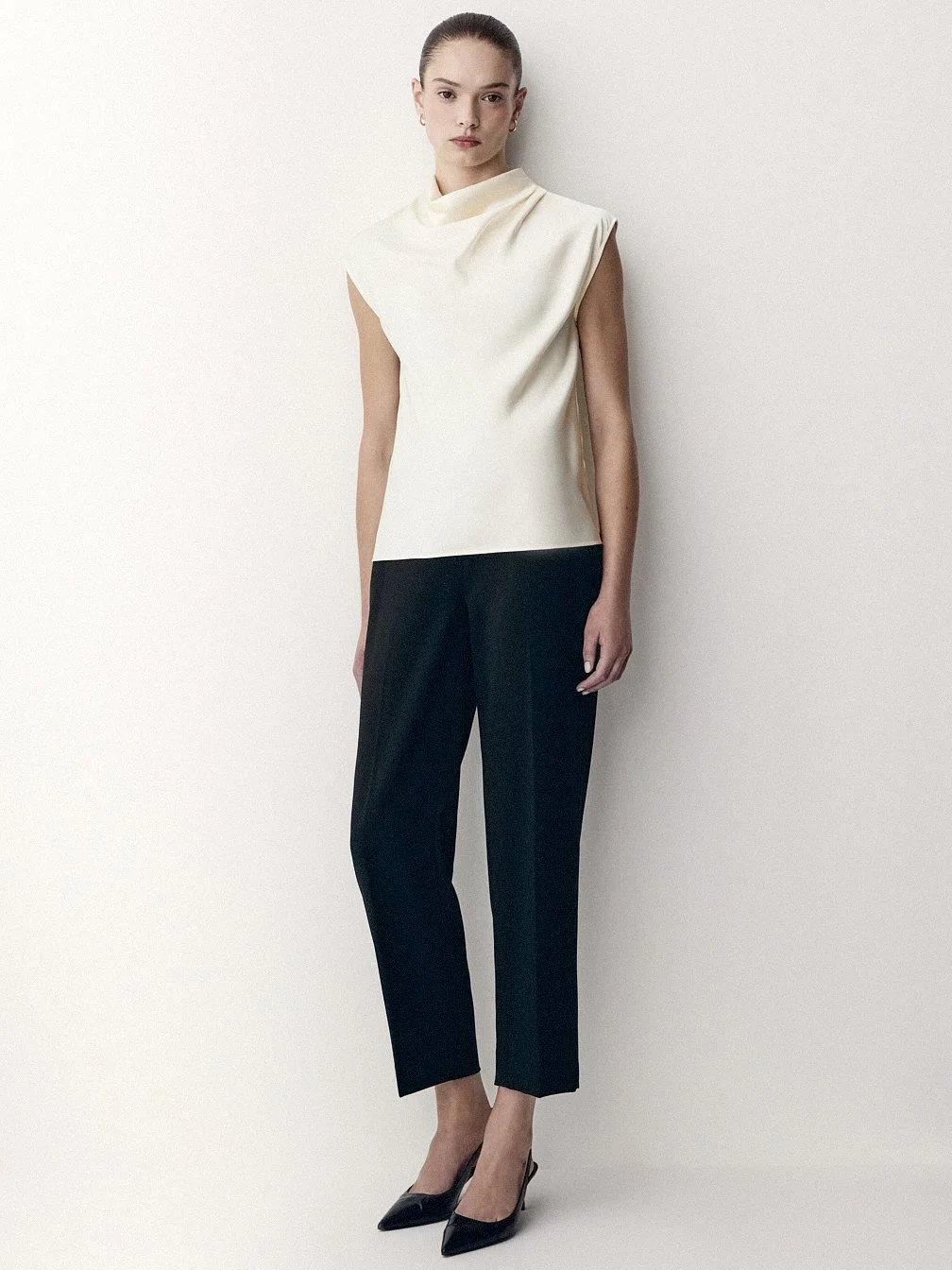 Woman in a sleeveless cream top, cropped black trousers and pointed black shoes.