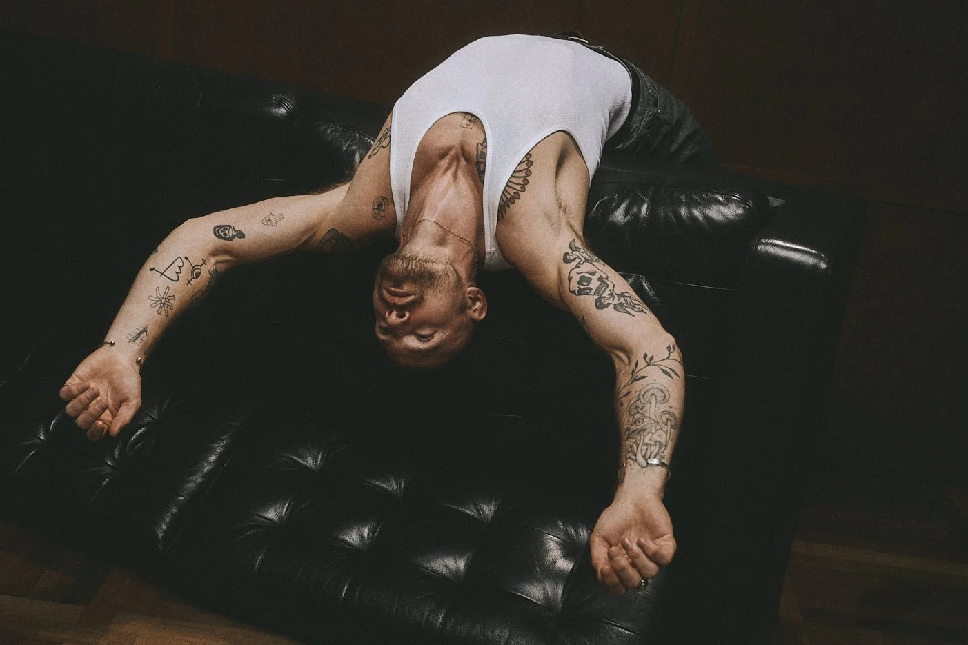 Tattooed man lying backwards over a leather sofa with his head and arms hanging down.