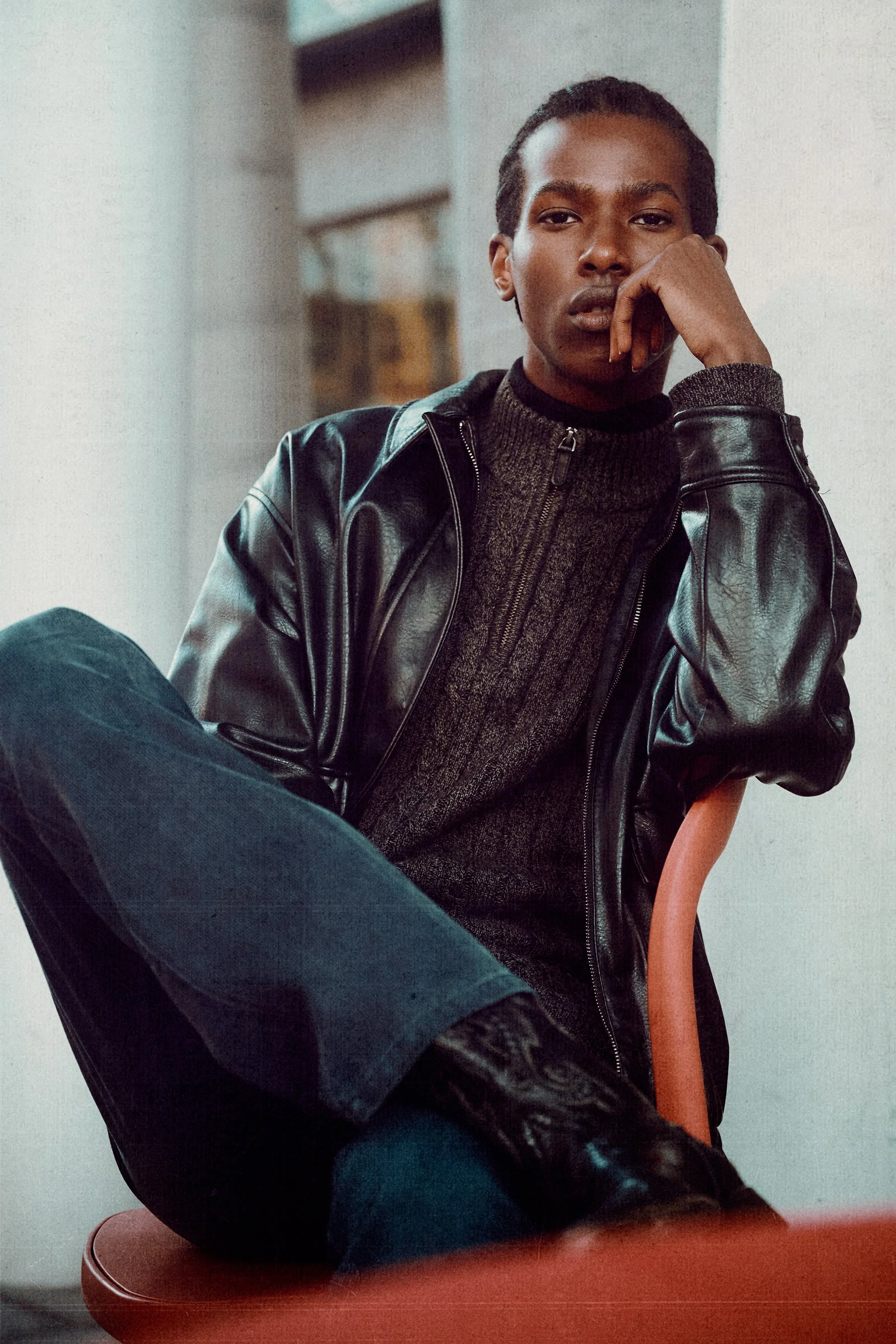 Man in a leather jacket and dark cable-knit sweater seated with his hand against his face.
