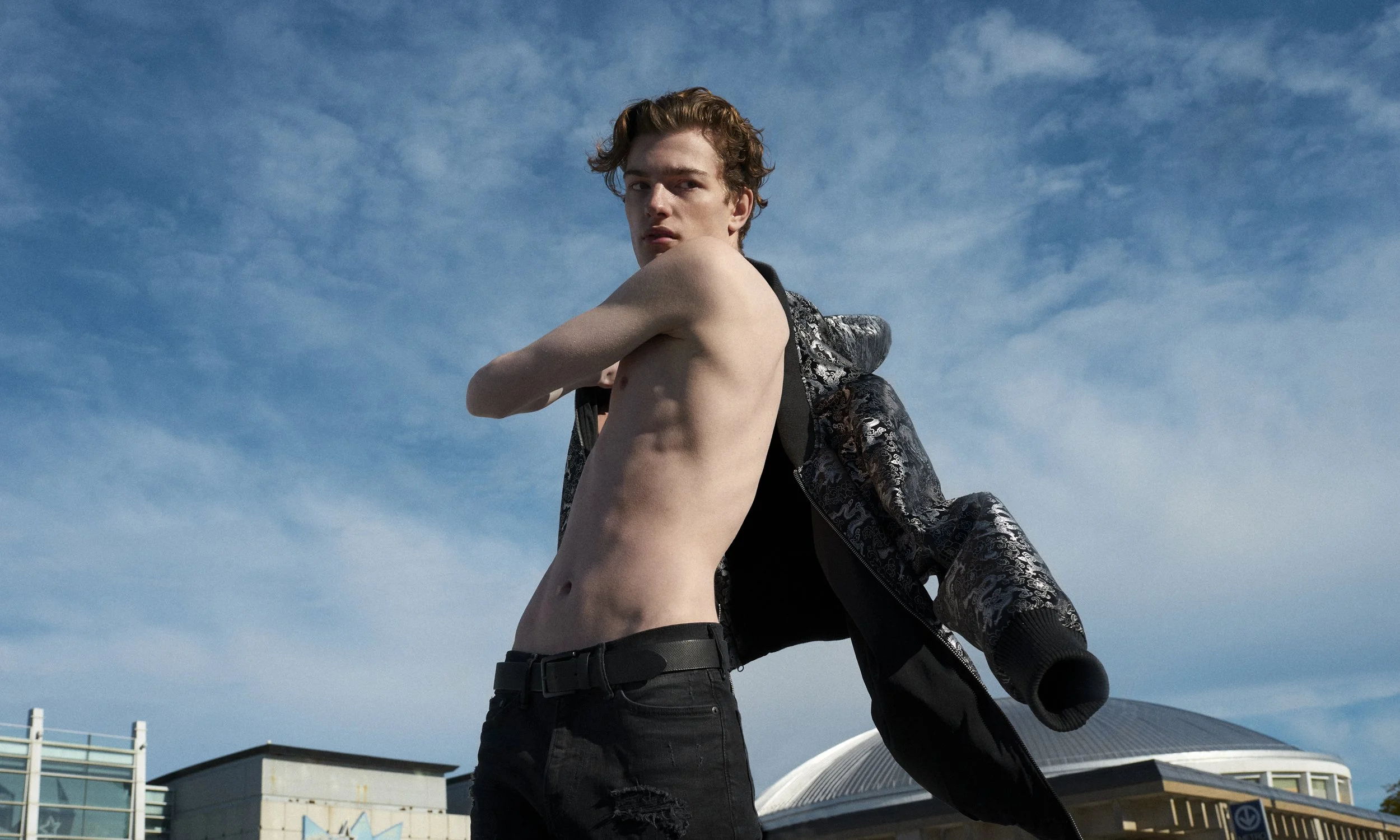 Shirtless man turning on a rooftop with a dark jacket swinging behind him beneath a blue sky.