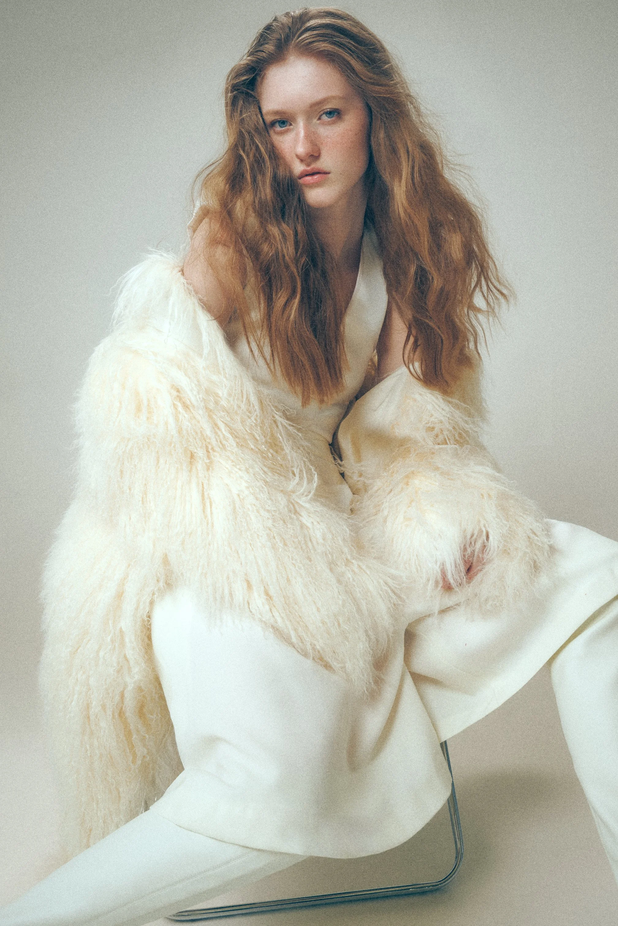 Woman seated in a cream outfit and fluffy white coat, with long loose hair.