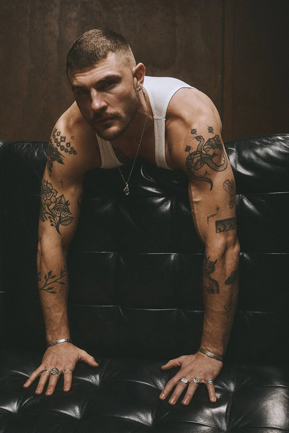 Tattooed man in a white tank top leaning forward on a leather sofa with both hands on the seat.