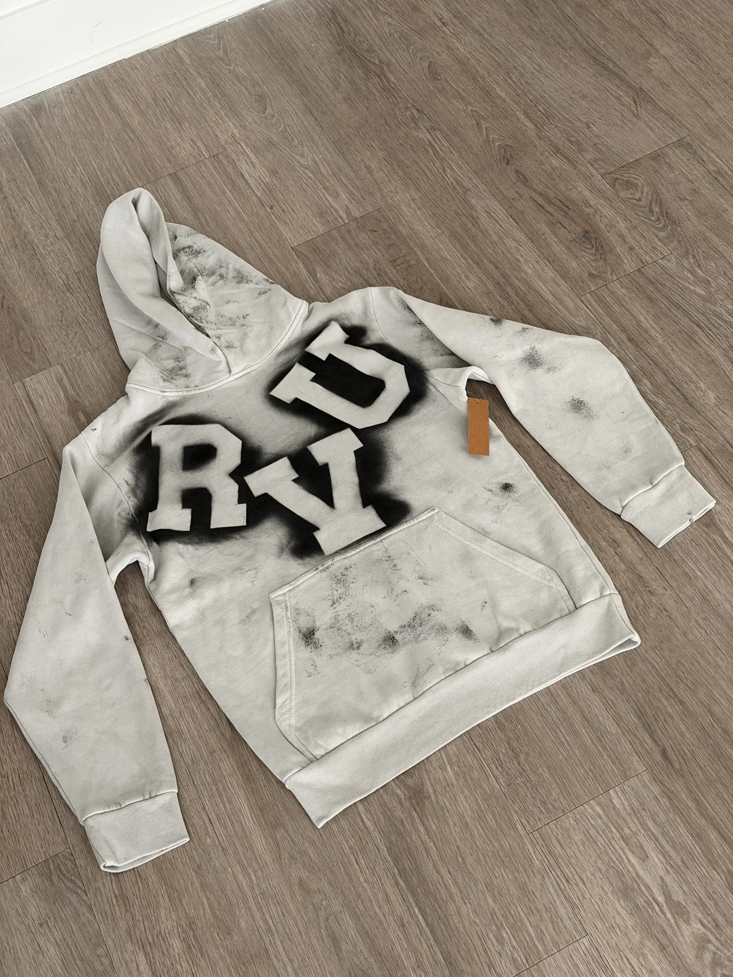 A white hoodie with black graffiti-style letters and black spray paint stains, lying on a wooden floor.