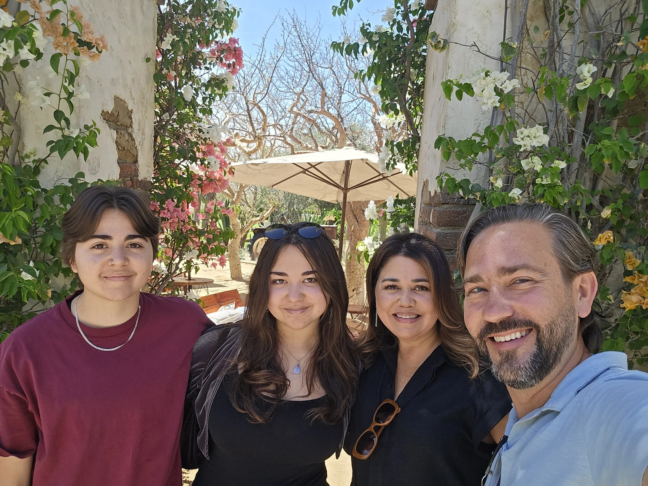 Four people smiling outdoors on a sunny day with greenery and flowers in the background.
