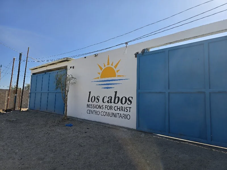 A white wall with a colorful logo of a sun setting over water, and the text 'los cabos' and 'MISSIONS FOR CHRIST' and 'CENTRO COMUNITARIO' underneath. Blue gate on the right side, trees and power lines in the background, under a clear blue sky.