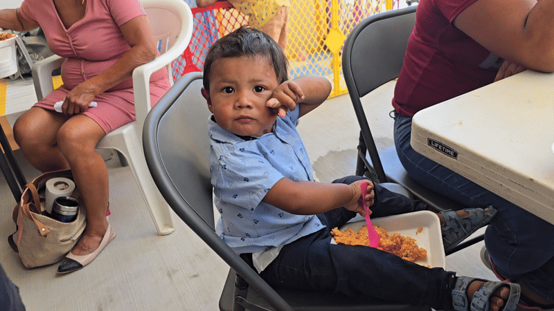 A young boy sitting in a high chair at a table, holding a pink spoon with food in a bowl, making a serious face while looking at the camera. An adult woman and man are partially visible nearby, with another woman sitting in the background.