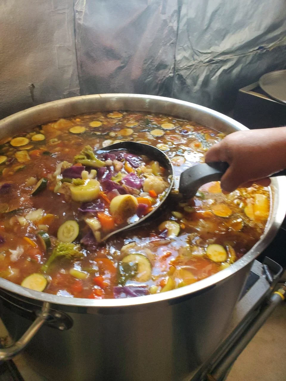 A large pot of vegetable soup being served with a ladle, filled with chopped vegetables like carrots, zucchini, and eggplant in a tomato broth.