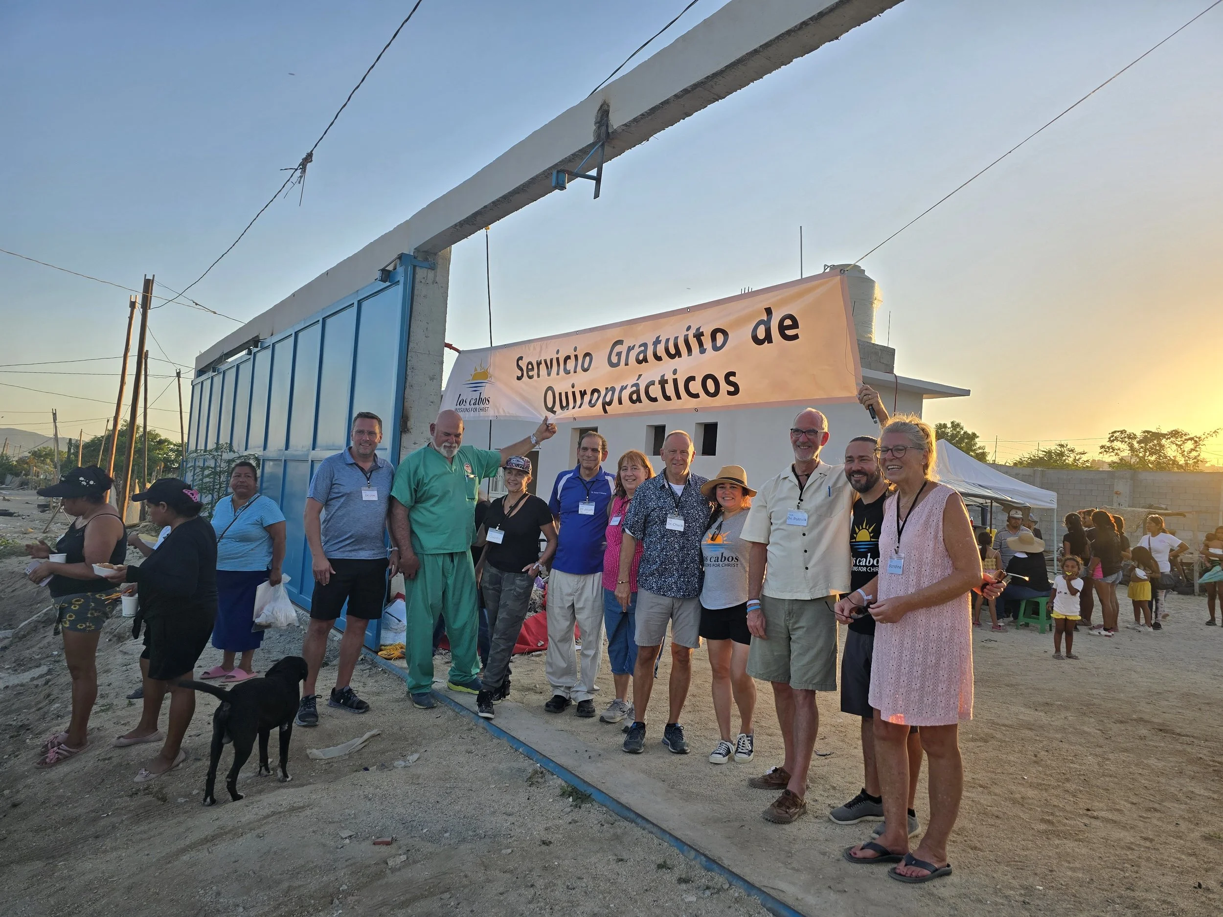 Group of people standing in front of a sign that reads 'Servicio Gratuito de Quiroprácticos' during sunset, some holding the sign and smiling, with a dirt area and people in the background.