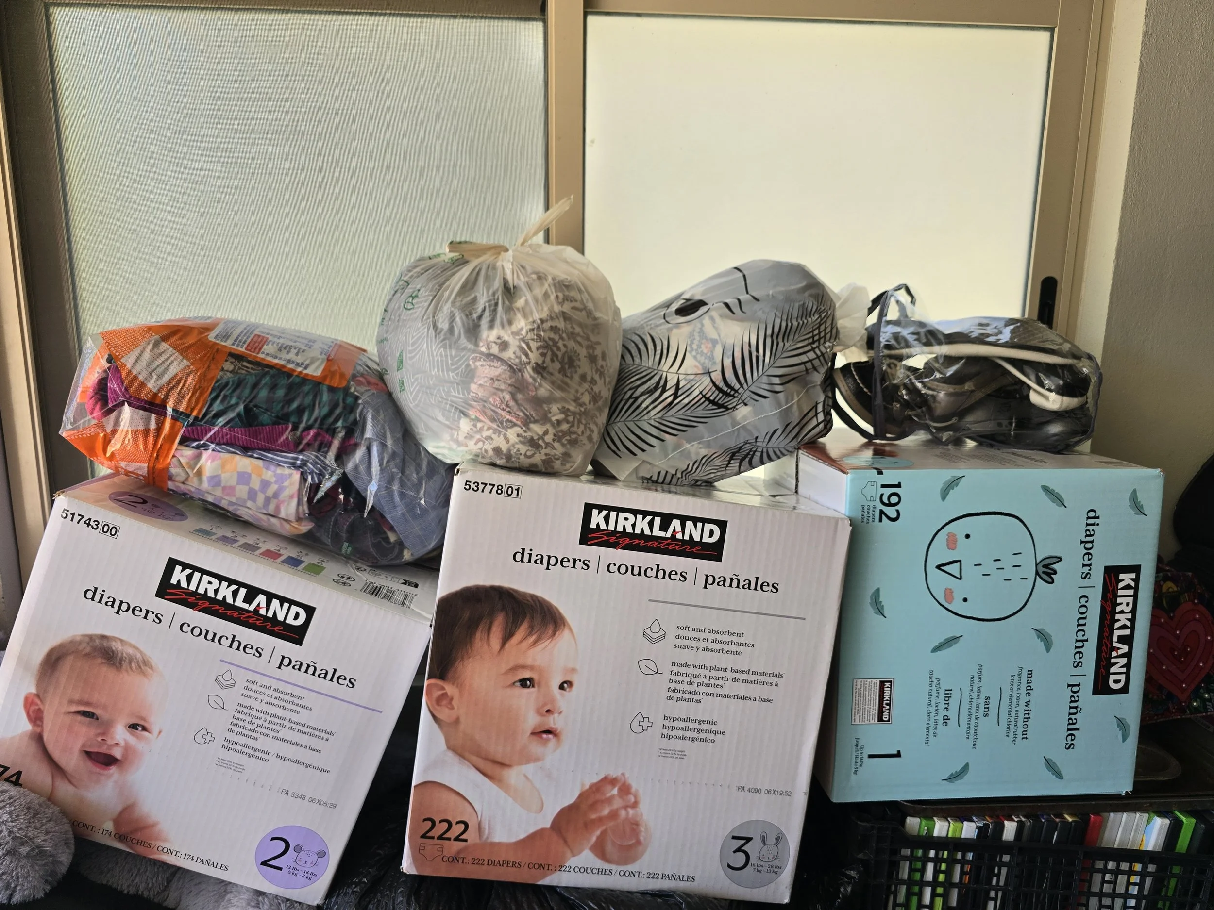 Boxes of Kirkland brand baby diapers with illustrations of babies and packaging details, along with bags of clothing or other items on top, in front of a window.