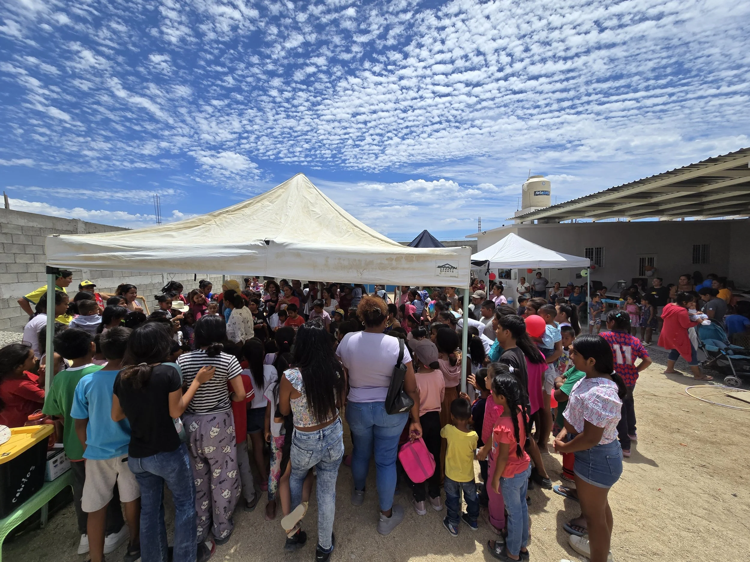 A large group of people, including children and adults, gathered outdoors under tents during a sunny day with a blue sky and scattered clouds. The scene appears to be at a community event or celebration.