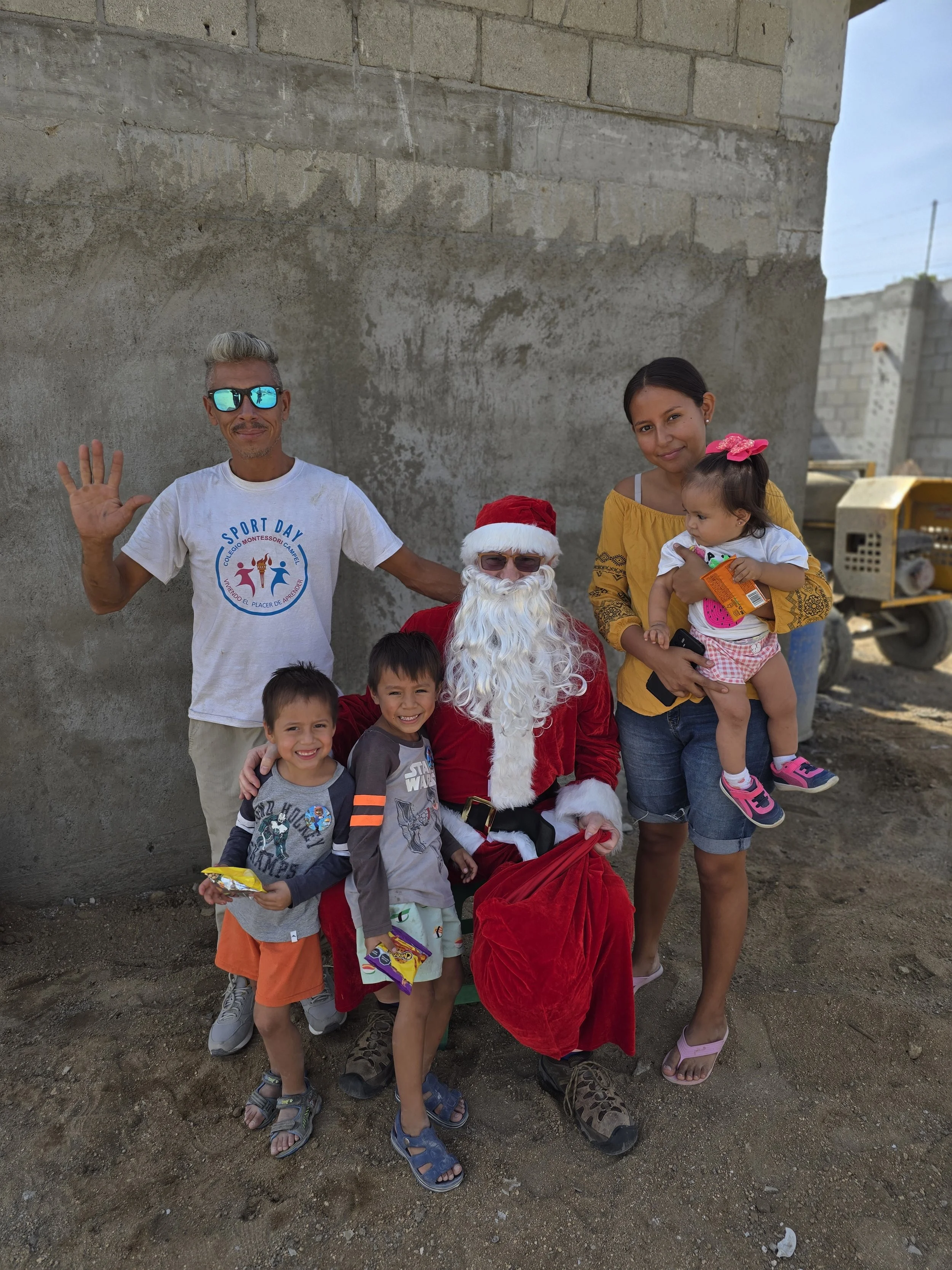 Family group with Santa Claus posing outdoors in front of a concrete wall and ground, holding candy and children, during daytime.
