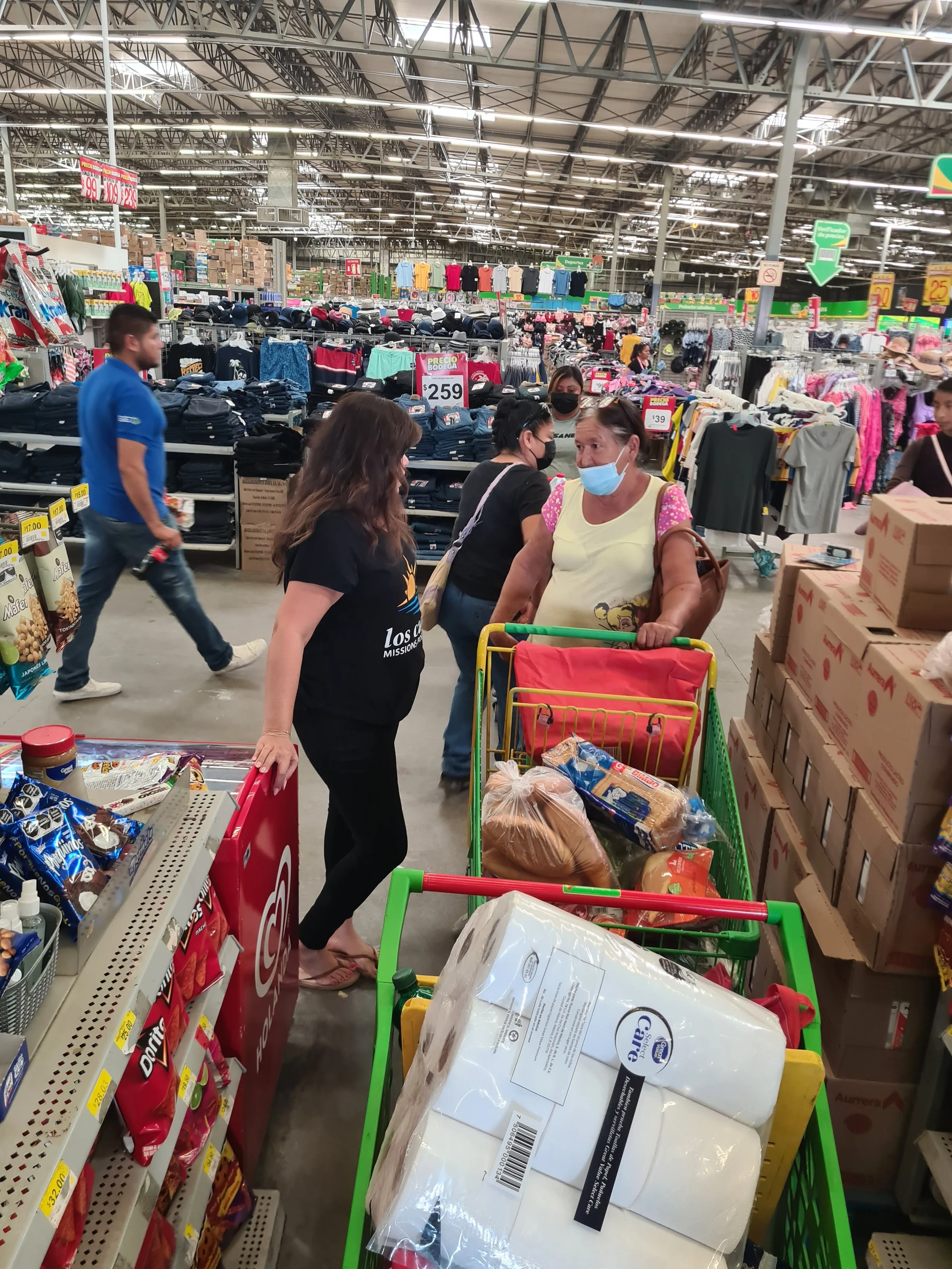 People shopping in a warehouse store aisle, with shopping carts filled with groceries and items, some wearing face masks.