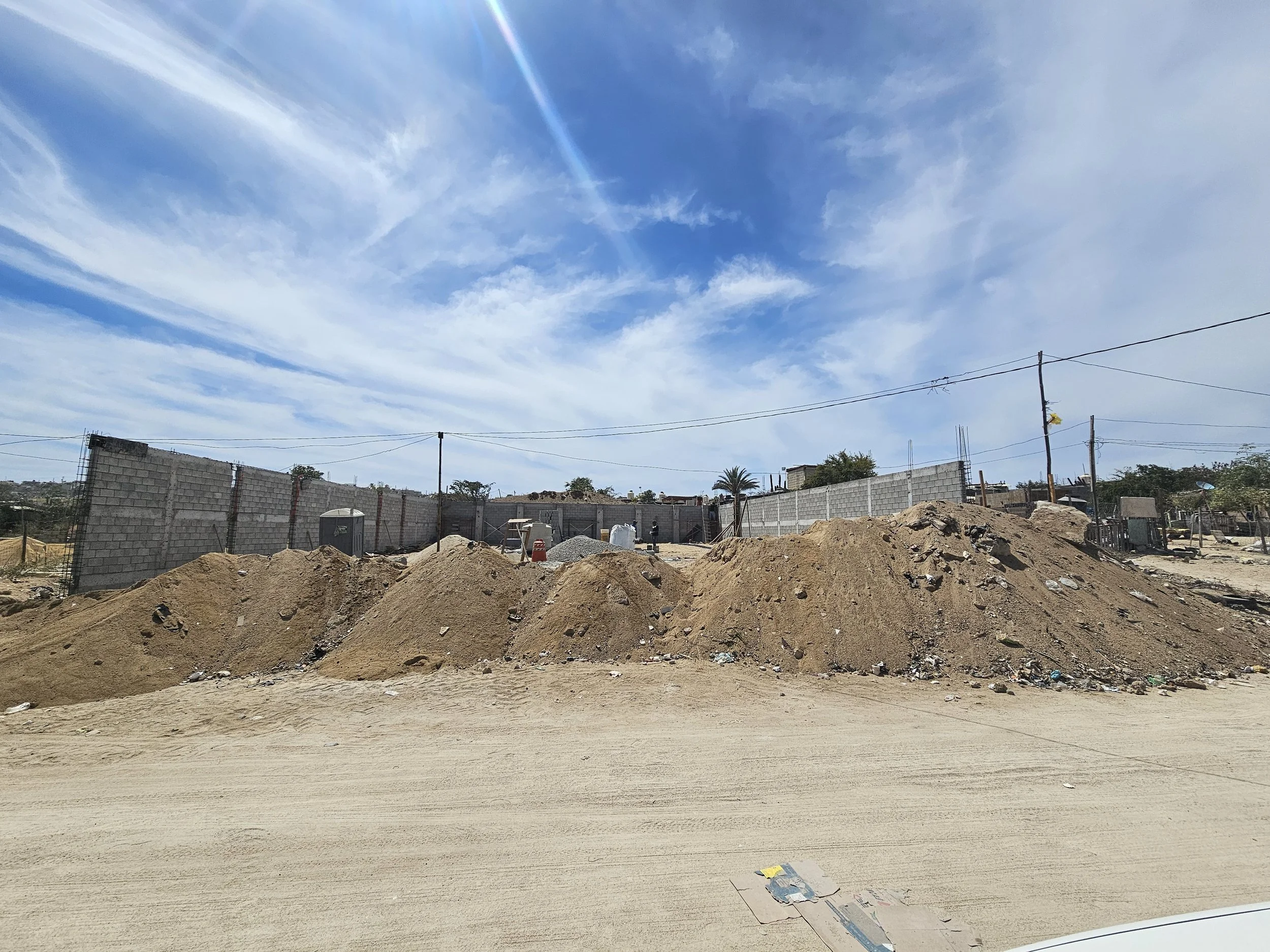 View of a construction site with dirt mounds, a partially built brick wall, utility poles, and a clear blue sky with scattered clouds.
