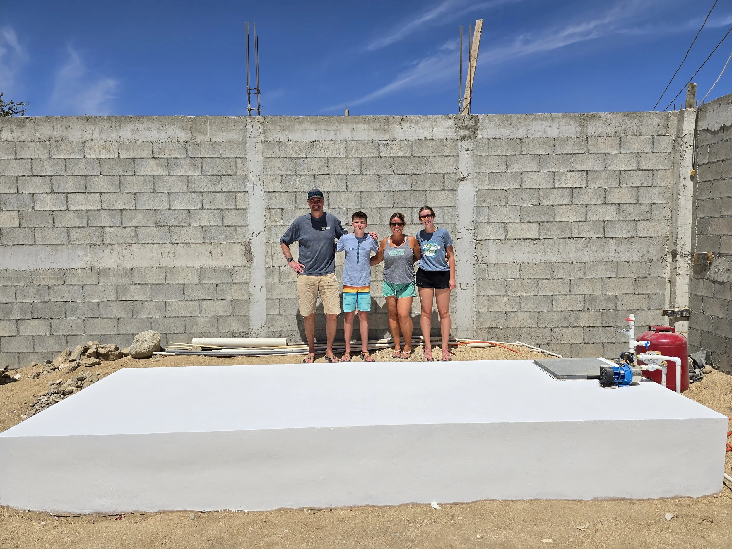 Four people standing on a freshly painted white platform outdoors, with a concrete block wall and blue sky in the background. The group includes two women, a boy, and a man, all smiling at the camera.