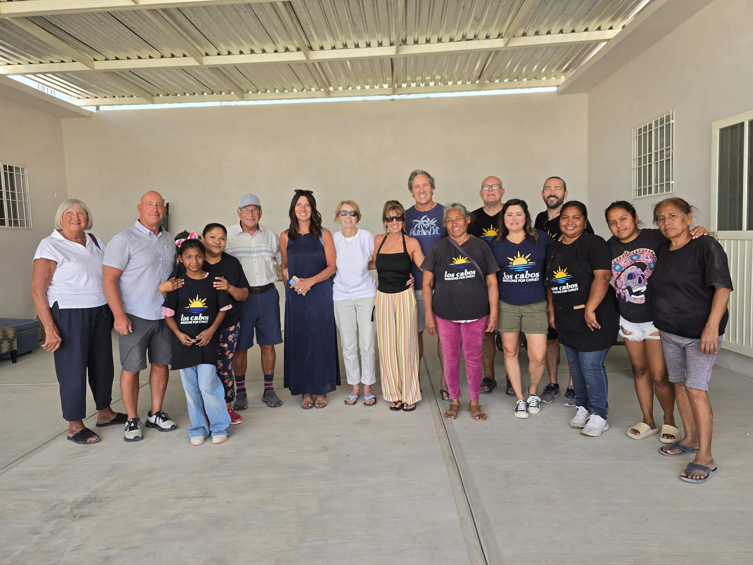 Group of diverse people gathering outdoors under a covered patio, some wearing matching black t-shirts with a yellow sun and the words 'Los Cabos' and 'Missions for Christ'.