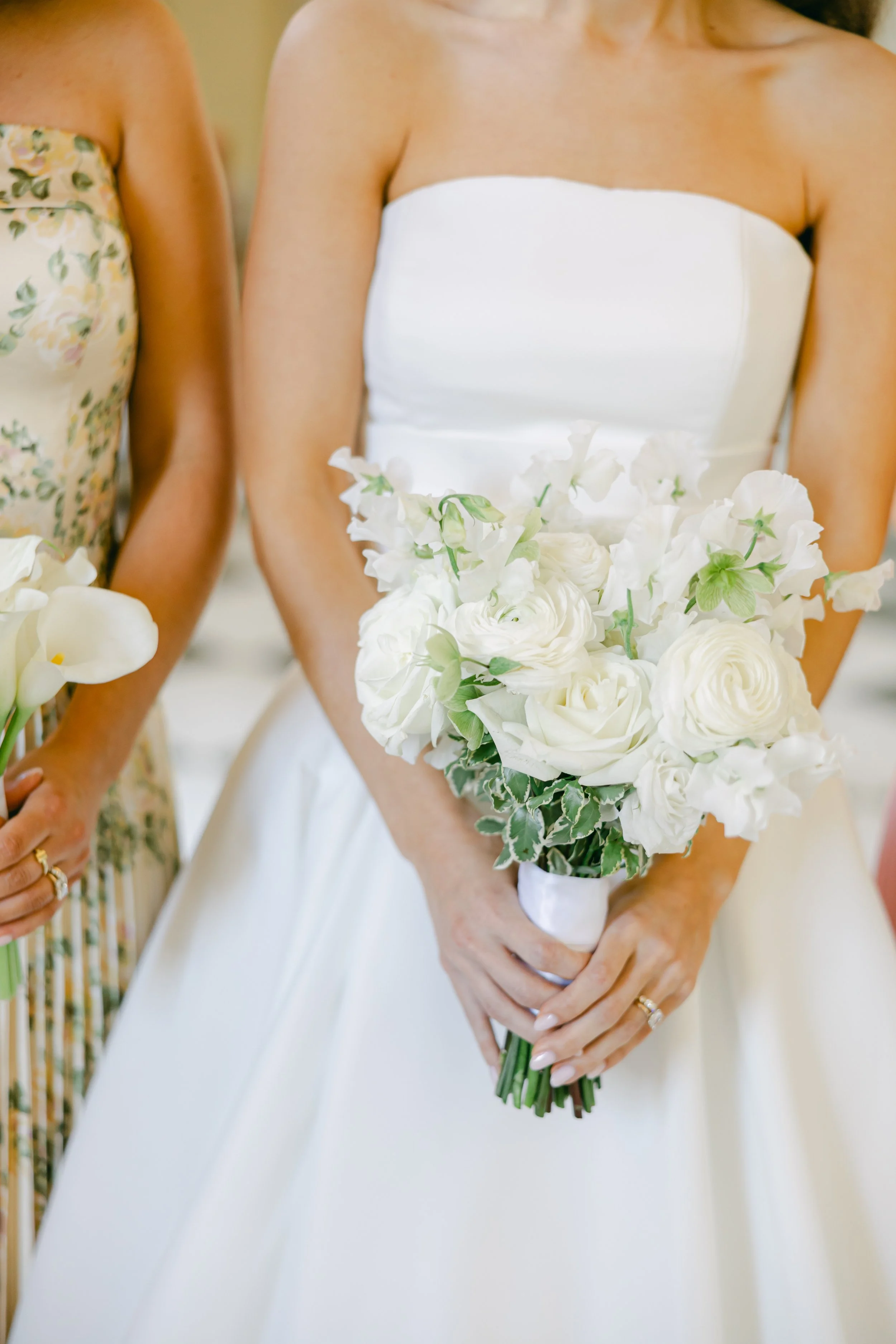 Close-up of a woman wearing a white strapless dress holding a bouquet of white flowers at a wedding, with another woman partially visible beside her.