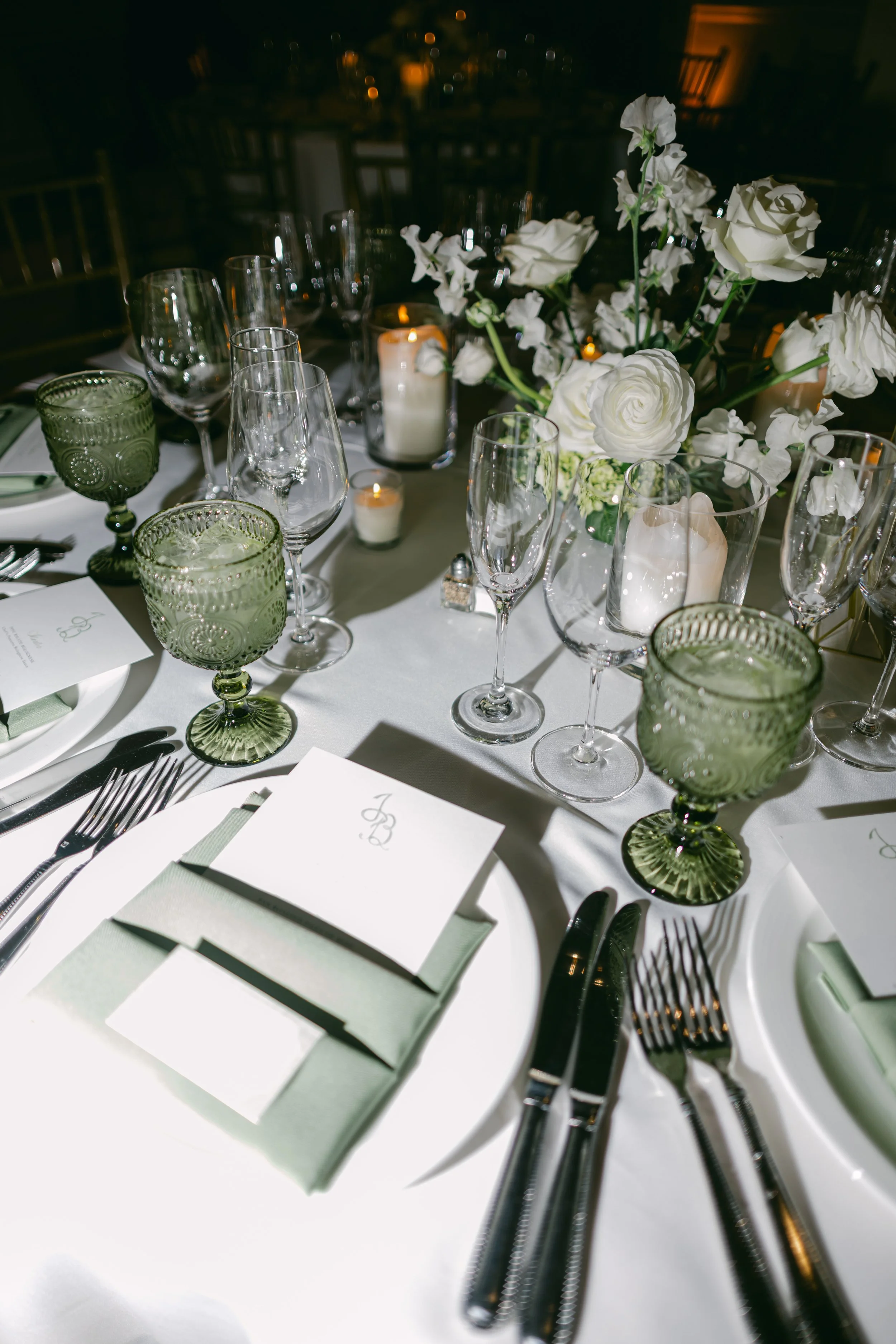 A formal dining table set for a special occasion with white tablecloth, assorted glassware including wine glasses and green-patterned goblets, silverware, white floral centerpiece, and candles in glass holders.