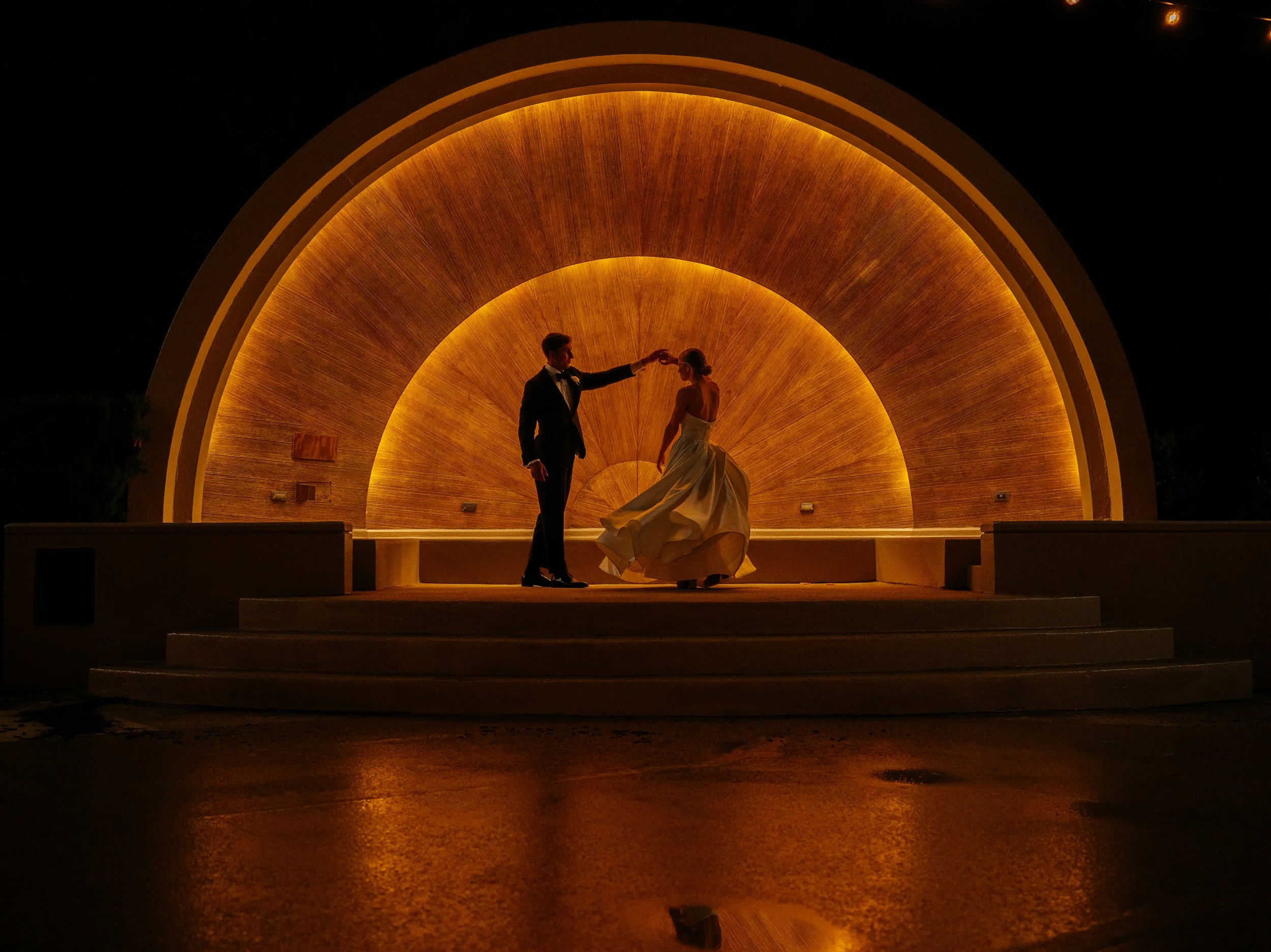 A couple in wedding attire dancing under a large, illuminated, semi-circular decorative structure at night.