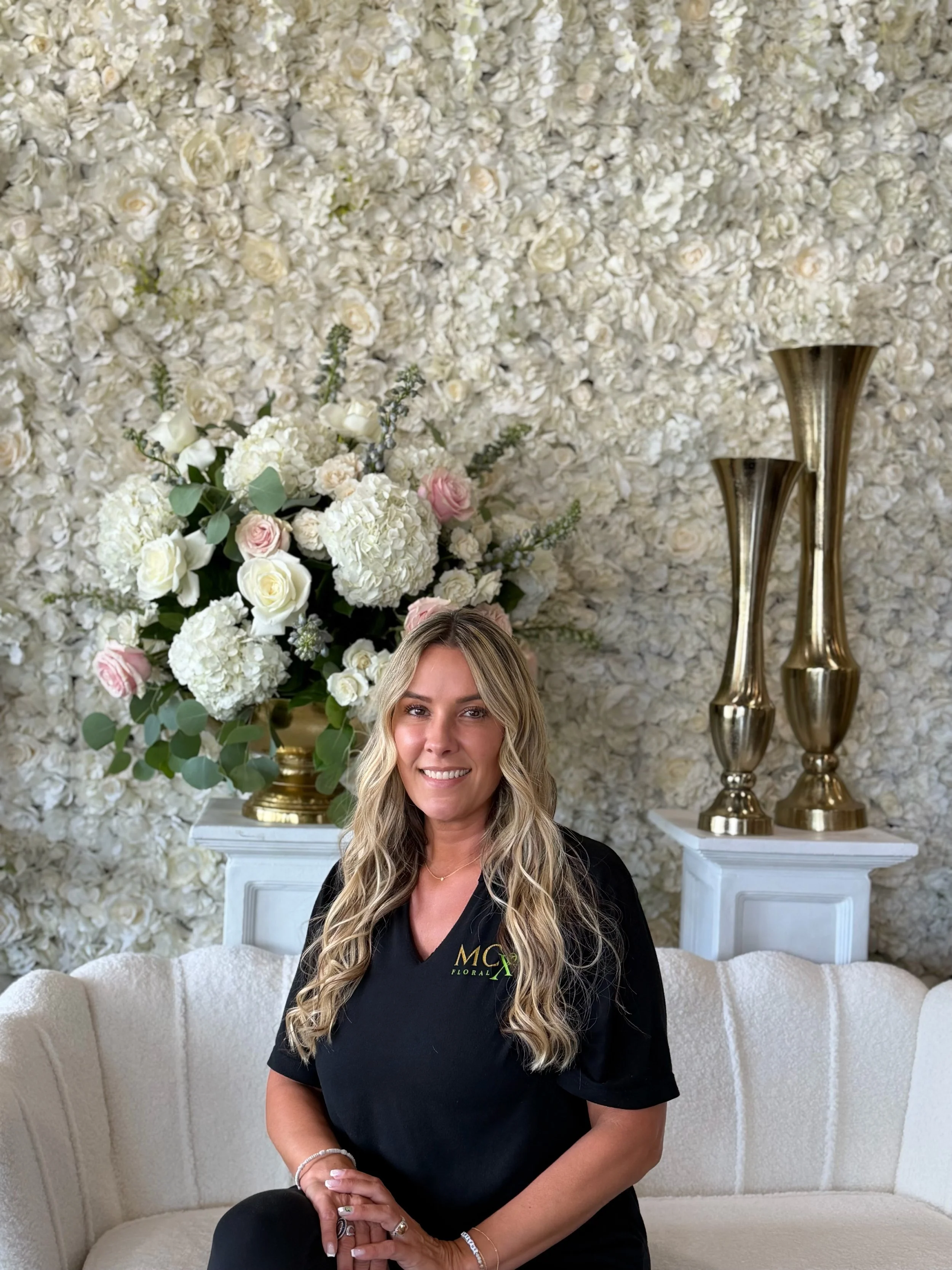 Monica Chimes is seated on a white couch. Behind her, there is a floral wall with white roses, hydrangeas, and pink roses, and two tall, gold decorative vases on white pedestals.